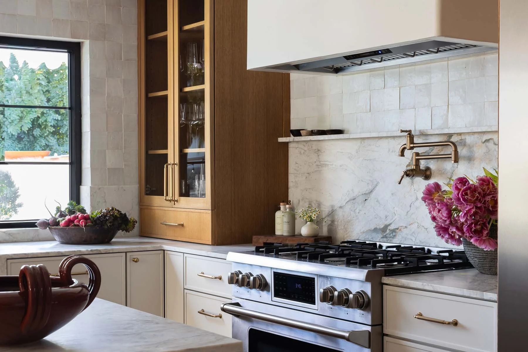 Kitchen with white marble countertops, beige cabinets, and a stainless steel stove. A window with greenery outside, a bowl of fruit on the counter, and flowers in a vase add decorative touches.