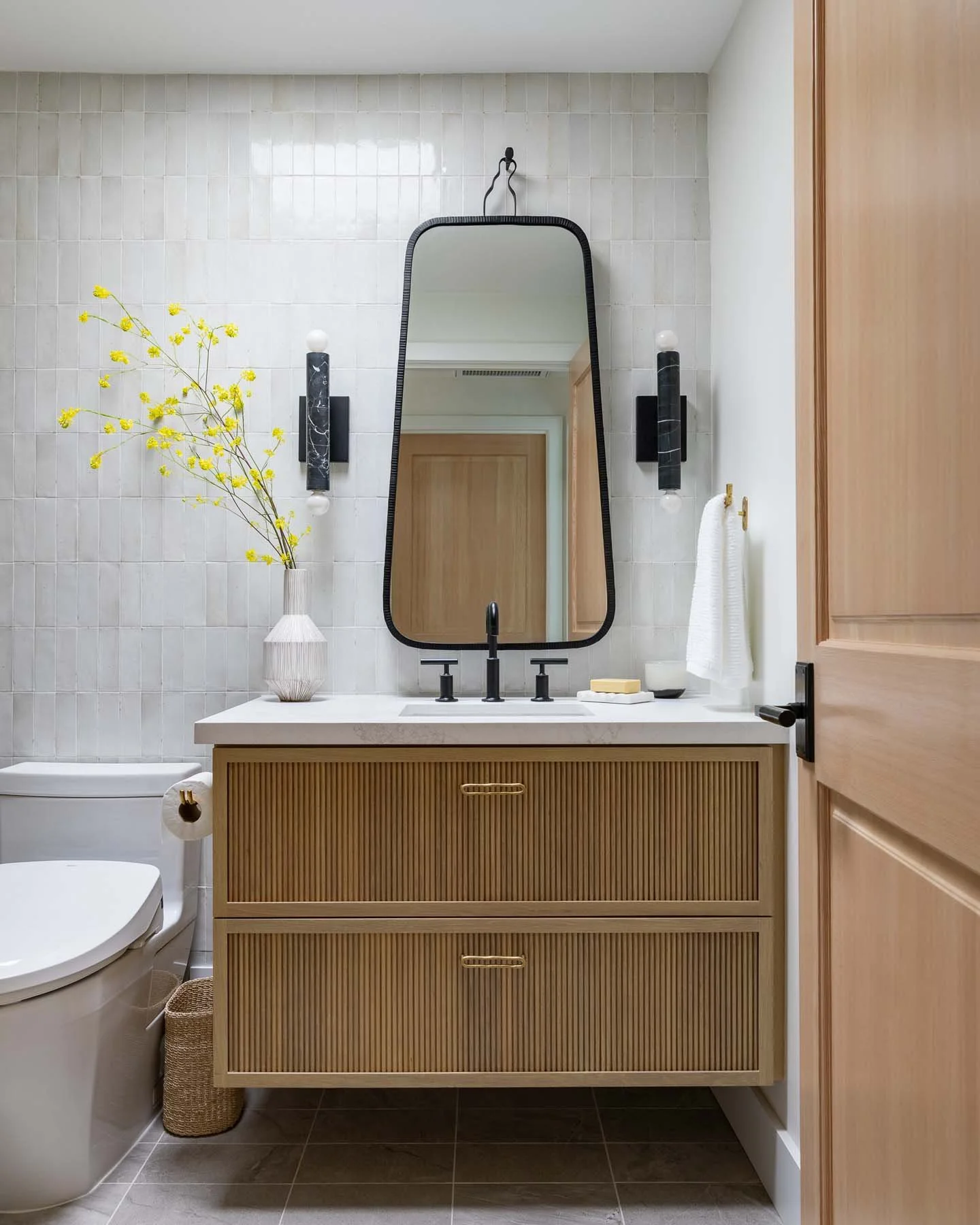Modern bathroom with a rectangular mirror, black light fixtures, a wooden vanity with ribbed doors, a white countertop, a vase with yellow flowers, a soap dish, and a towel hanging on a hook.