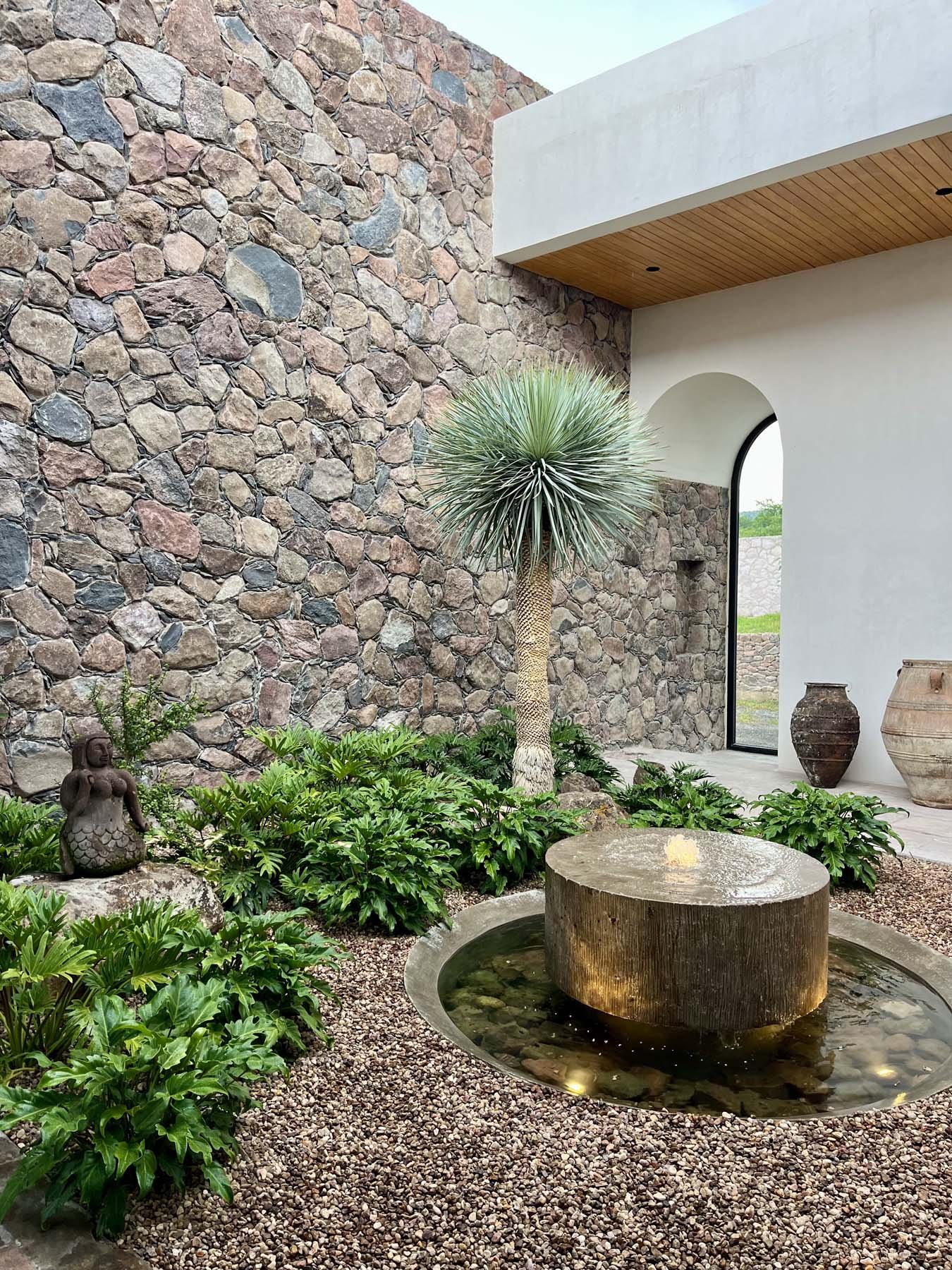 A modern courtyard featuring a stone wall with multicolored rocks, a small water fountain with a stone base, lush green plants including a large spiky desert plant, and decorative pottery. There is an arched doorway with a white wall and wooden ceiling in the background.
