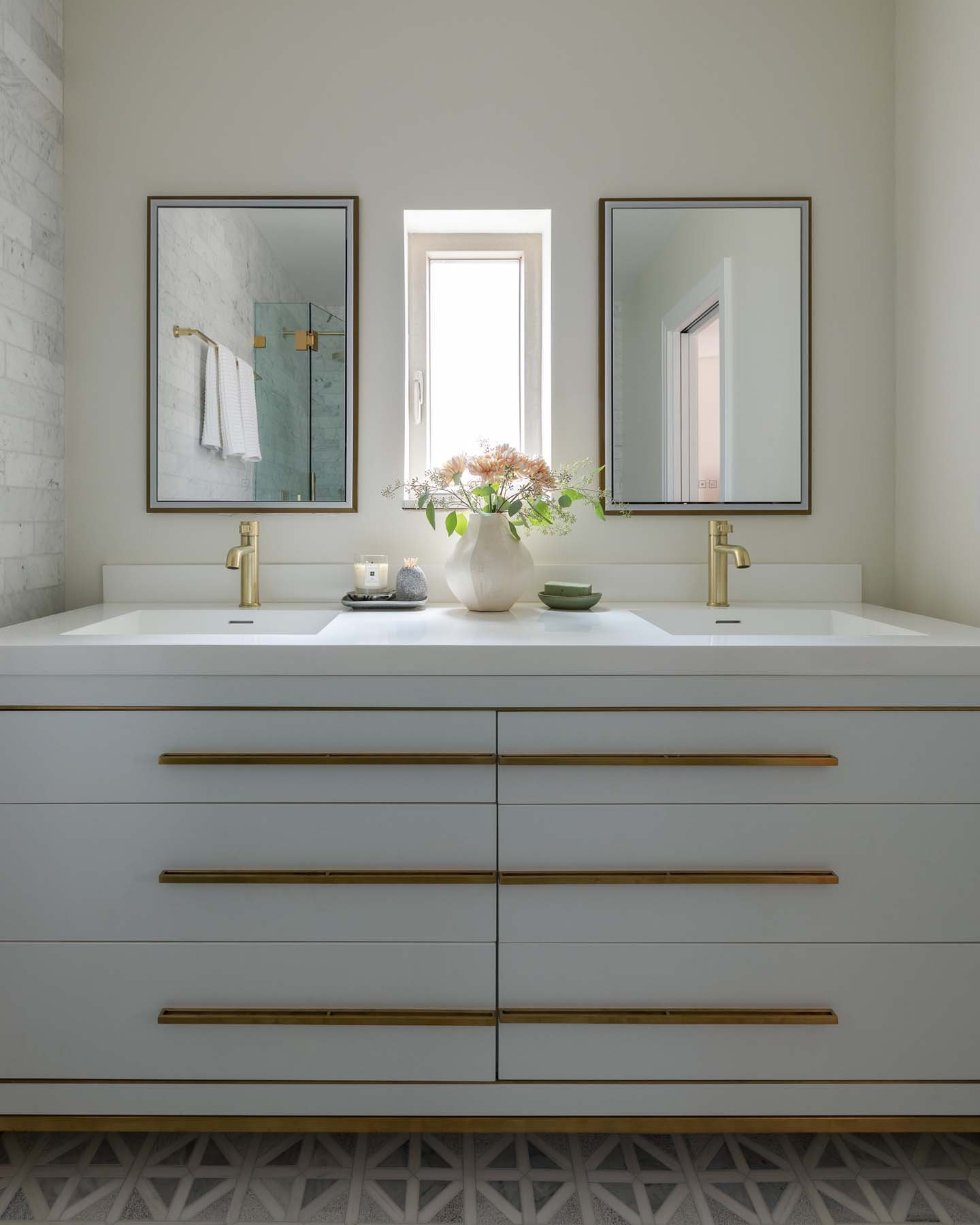 Bathroom vanity with two sinks, gold faucets, a white countertop, and a large vase with pink flowers between two mirrors. A window is centered above the vanity, and a shower with glass door and white towels is visible through the left mirror.