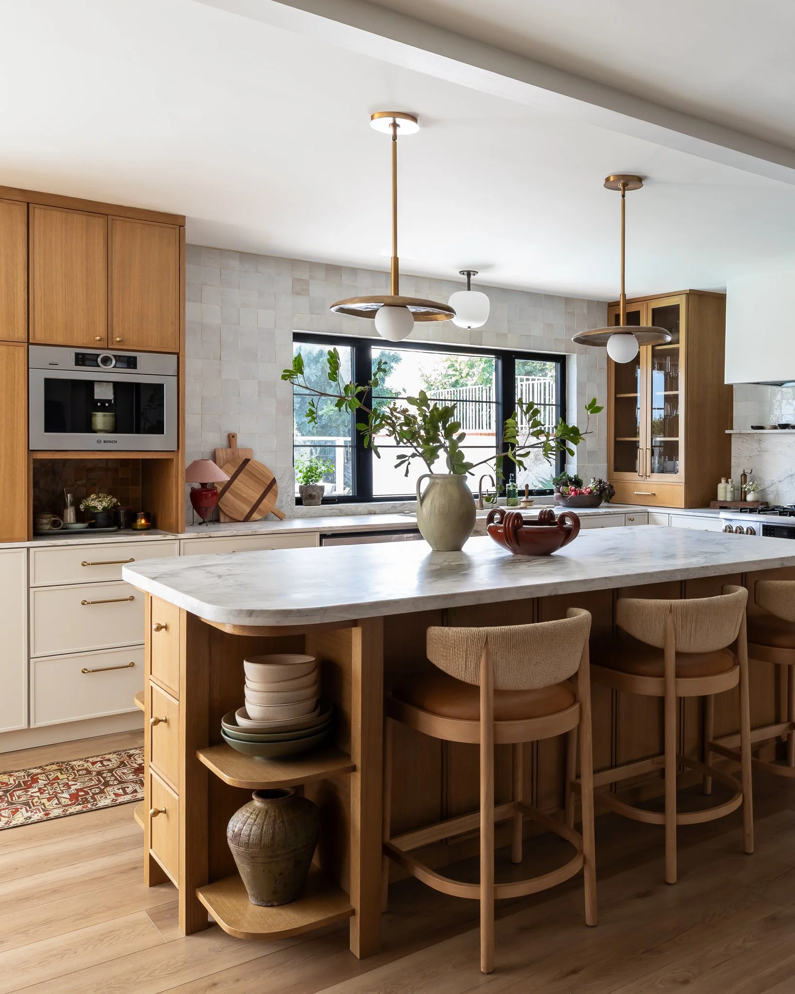 Modern kitchen with a white marble island counter, wood cabinetry, and pendant lights hanging from the ceiling. Plants and bowls are placed on the island, and large windows provide natural light.