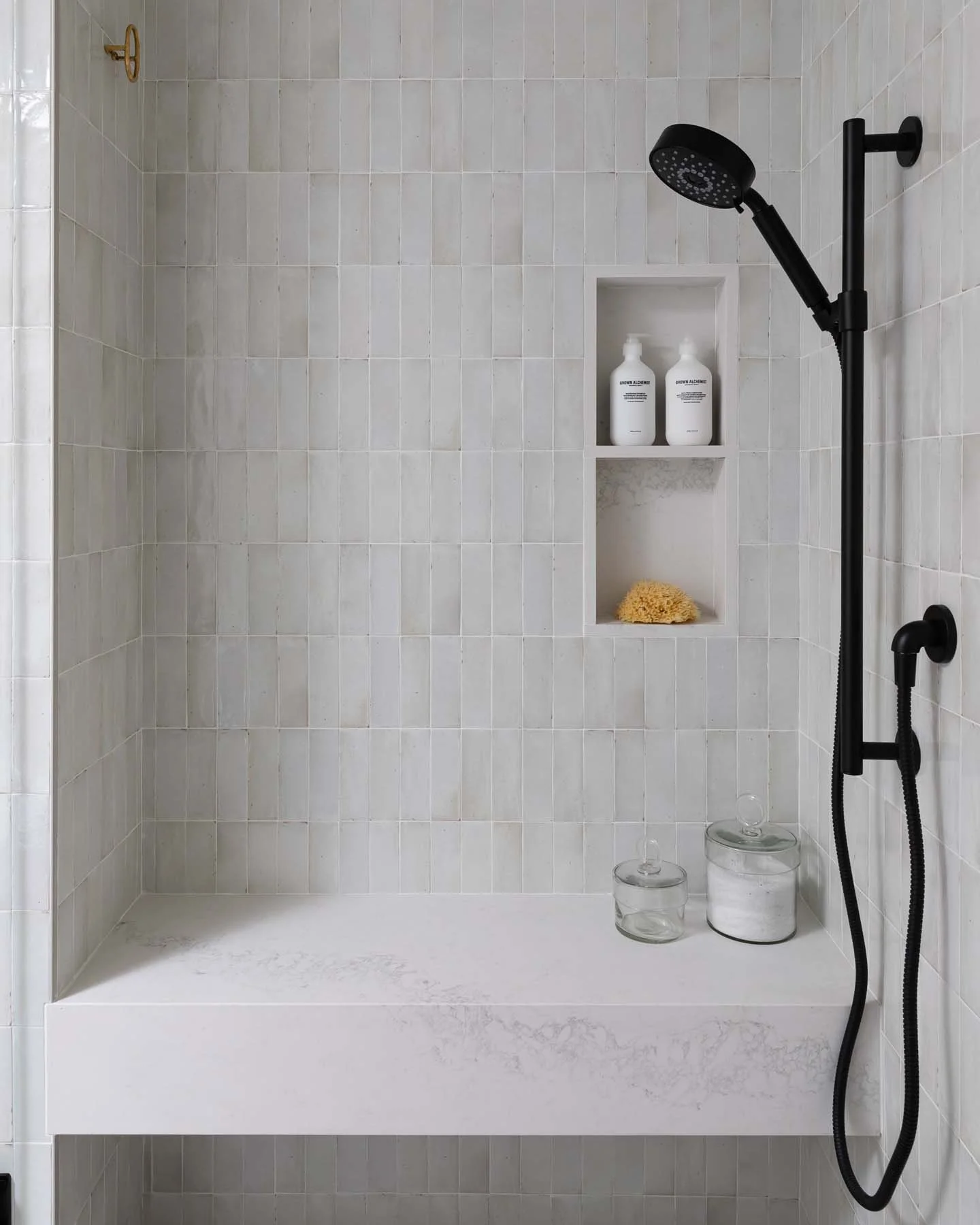 Empty shower with beige tiles, a black handheld shower head on a rail, built-in wall niche with two white bottles and a yellow sponge, and glass jars on the white shower bench.