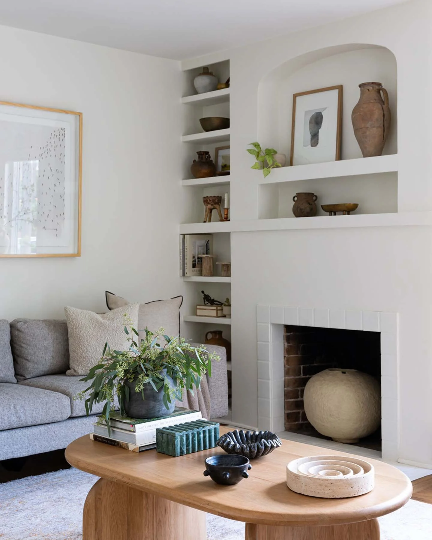 Living room with a light gray sofa, beige pillows, and a coffee table with decorative objects, a fireplace with a large round ceramic vase inside, and built-in white shelves with pottery and framed artwork.