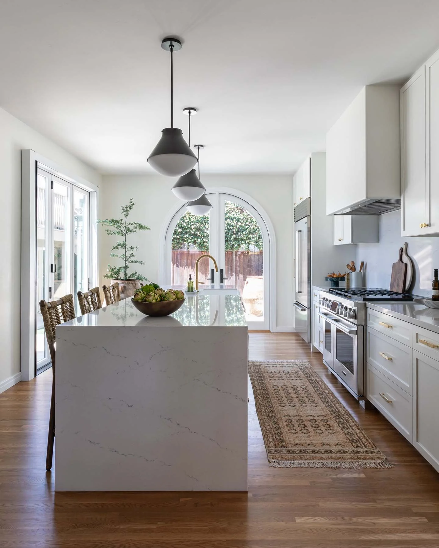 A bright, modern kitchen with white cabinets, a marble island with chairs, and a large window with an arched top showing outdoors greenery. There are black pendant lights hanging from the ceiling and a wooden floor.