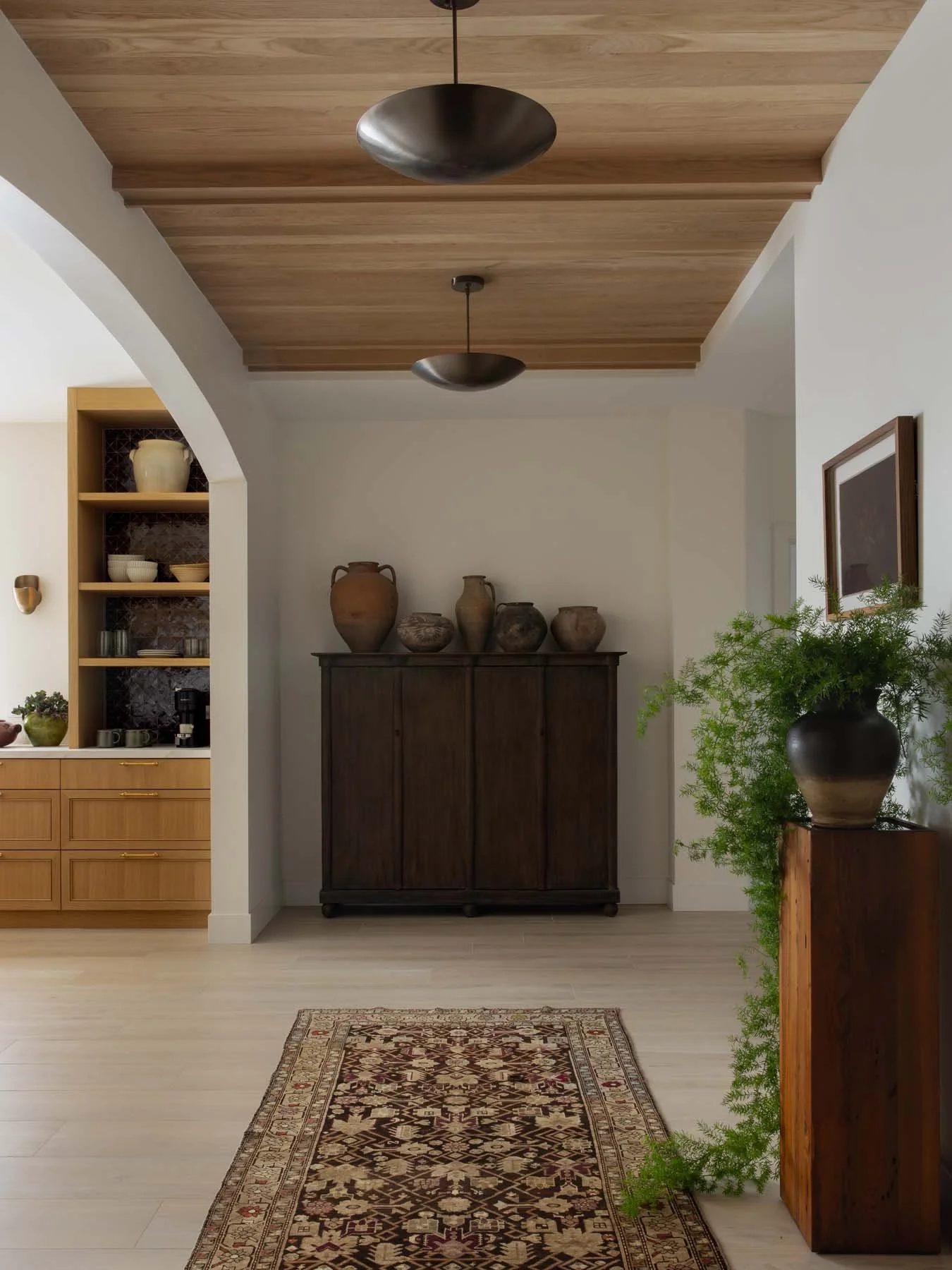 A hallway with wooden ceiling and floor, featuring a Persian-style rug, a dark wooden cabinet with decorative vases on top, and a framed picture on the wall. To the right, there is a green potted plant on a wooden stand.