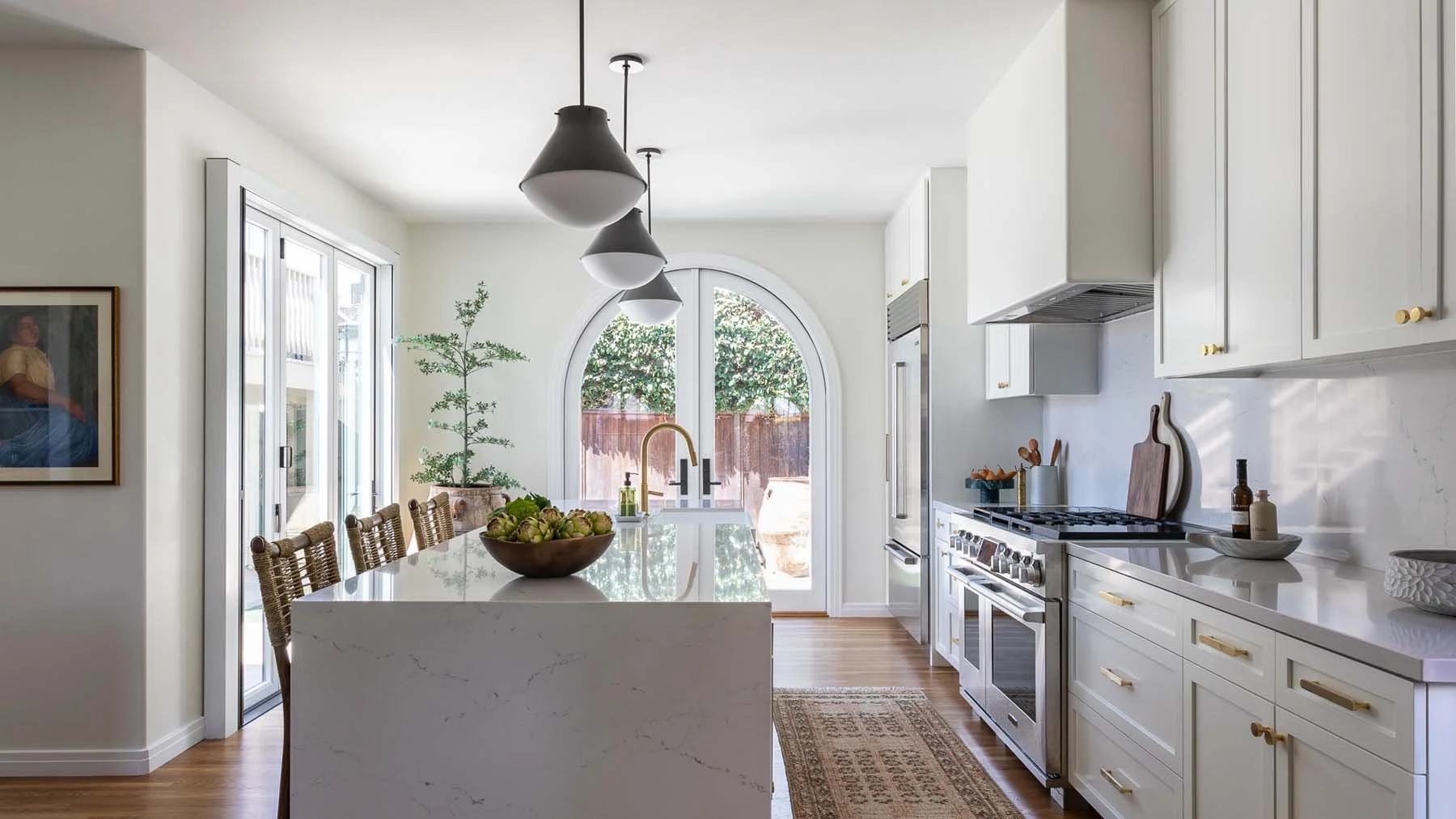 Bright modern kitchen with white cabinets, marble countertops, a kitchen island with a bowl of green apples, and a large arched window with a view of trees outside. There are black pendant lights hanging from the ceiling and a wooden dining table with woven chairs nearby.