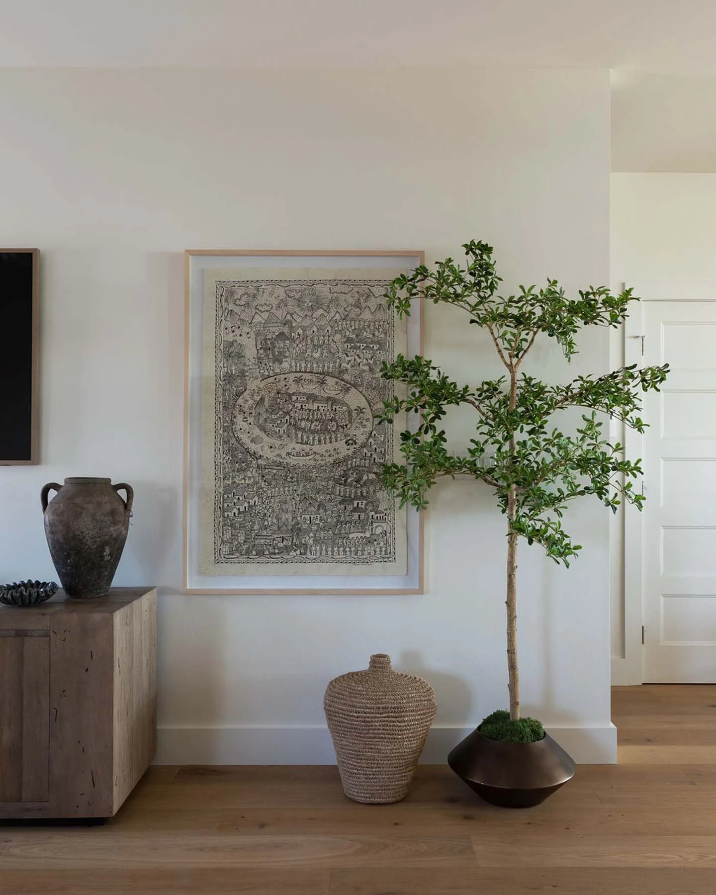 Interior of a living space featuring a tall potted tree, a framed black-and-white artwork, a wooden cabinet with a ceramic vase, and a woven basket on a wooden floor.