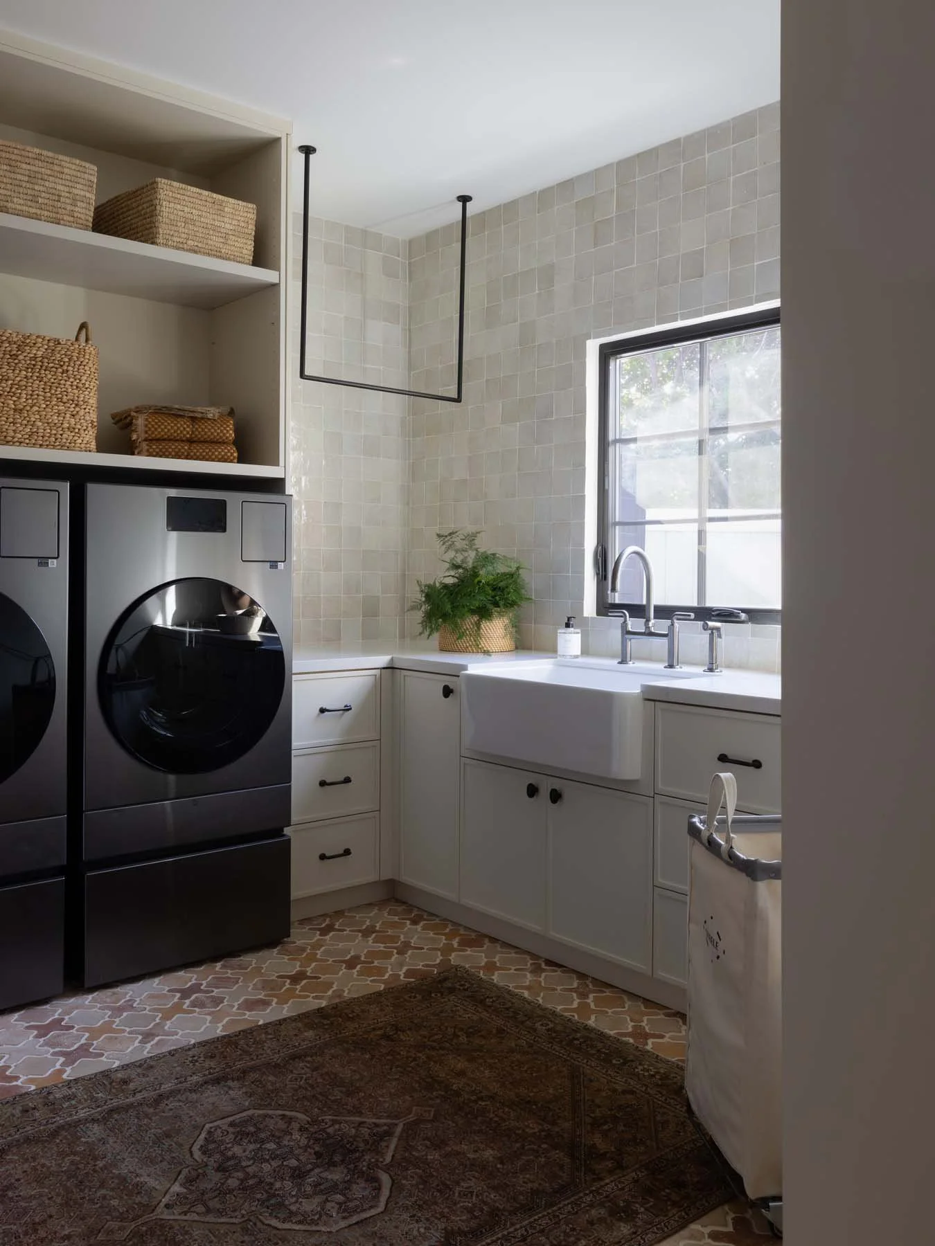 Laundry room with white cabinetry, a farmhouse sink under a window, two stainless steel washers and baskets on open shelves, a potted plant, and a patterned rug on the floor.