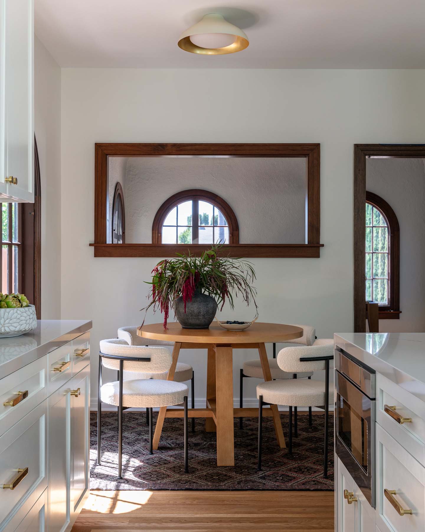A dining area with a round wooden table surrounded by four cream-colored chairs, a potted plant in a black vase on the table, and two arched windows with wooden frames in the background. White cabinetry with gold handles is visible on the sides, and a ceiling light fixture hangs above.