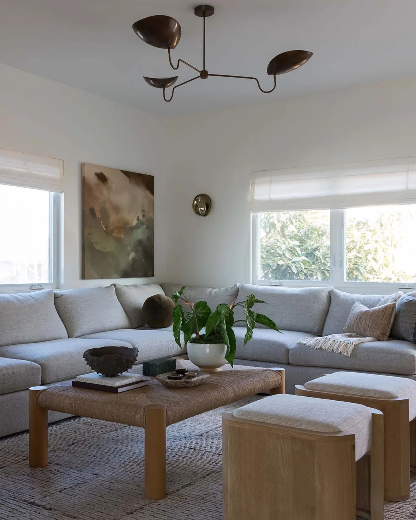 Bright living room with a large white sectional sofa, a wooden coffee table with decorative items, and two small stools. Large windows with white blinds allow natural light to fill the space, which is decorated with abstract art on the wall and a ceiling light fixture.