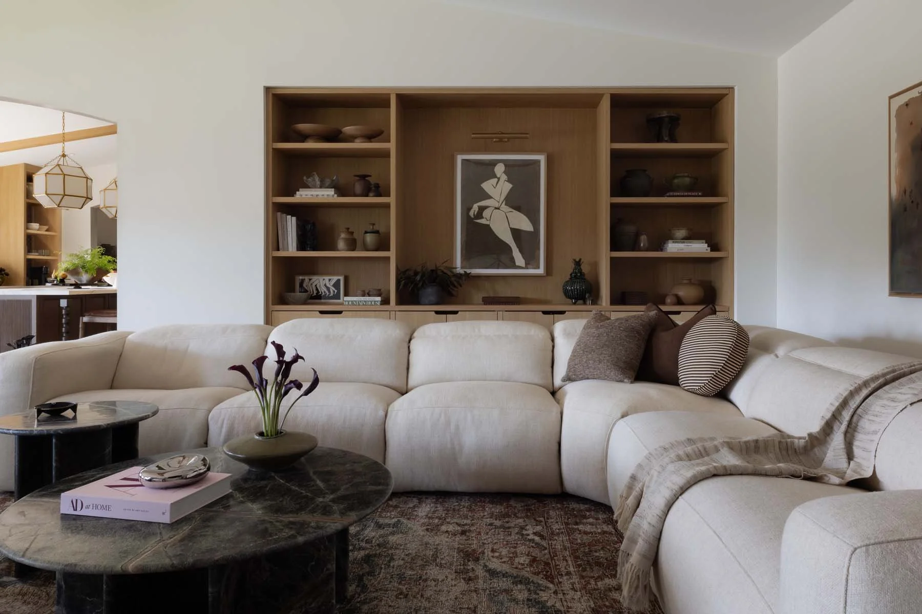 A cozy living room with a white sectional sofa adorned with pillows, a dark marble coffee table with a flower vase and books, and a built-in wooden bookshelf on the wall displaying decorative items and books. A framed abstract art piece hangs on the bookshelf, and a beige throw blanket is draped over the edge of the sofa.