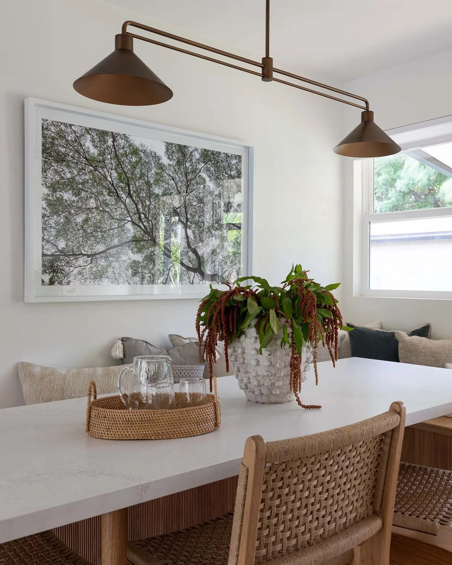 A white dining table with a large white textured vase holding a green and red plant at the center. Behind the table are woven rattan chairs and beige cushions. On the wall, there is a framed photograph of tree branches and leaves. There's a window on the right side letting in natural light, and a bronze-colored modern light fixture hanging from the ceiling.