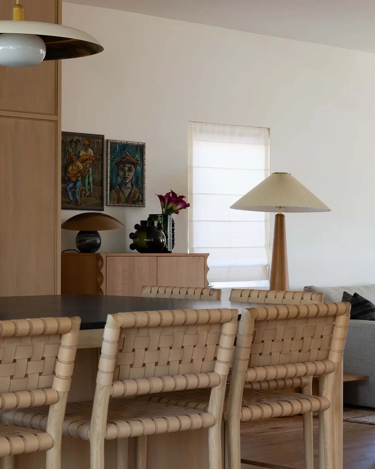 A dining room with a dark wood table, beige woven chairs, a wooden sideboard decorated with plants and lamps, a window with a white shade, and artwork on the wall.