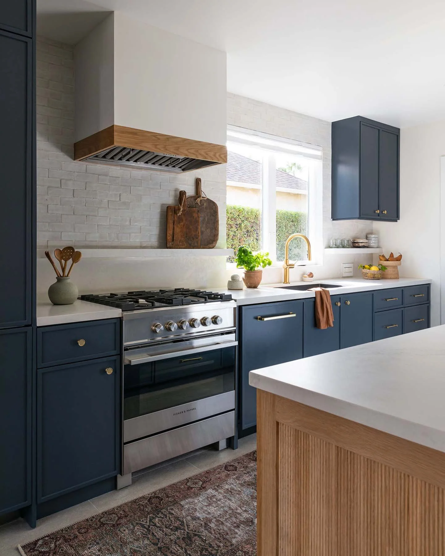 Modern kitchen with blue cabinets, white countertop, bronze hardware, stainless steel stove, backsplash with white brick tiles, window with greenery outside, potted plant, wooden cutting boards, and decorative items.