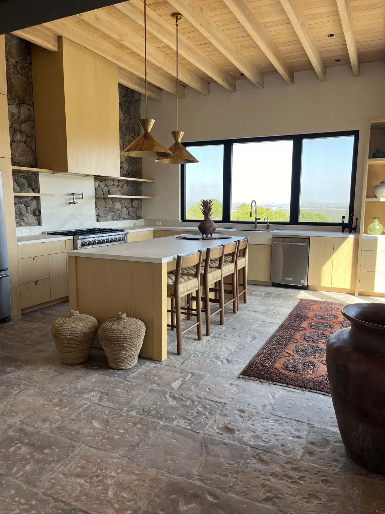 Modern kitchen with wooden ceiling, stone and wood walls, large window with a scenic view, kitchen island, four chairs, and decorative baskets.