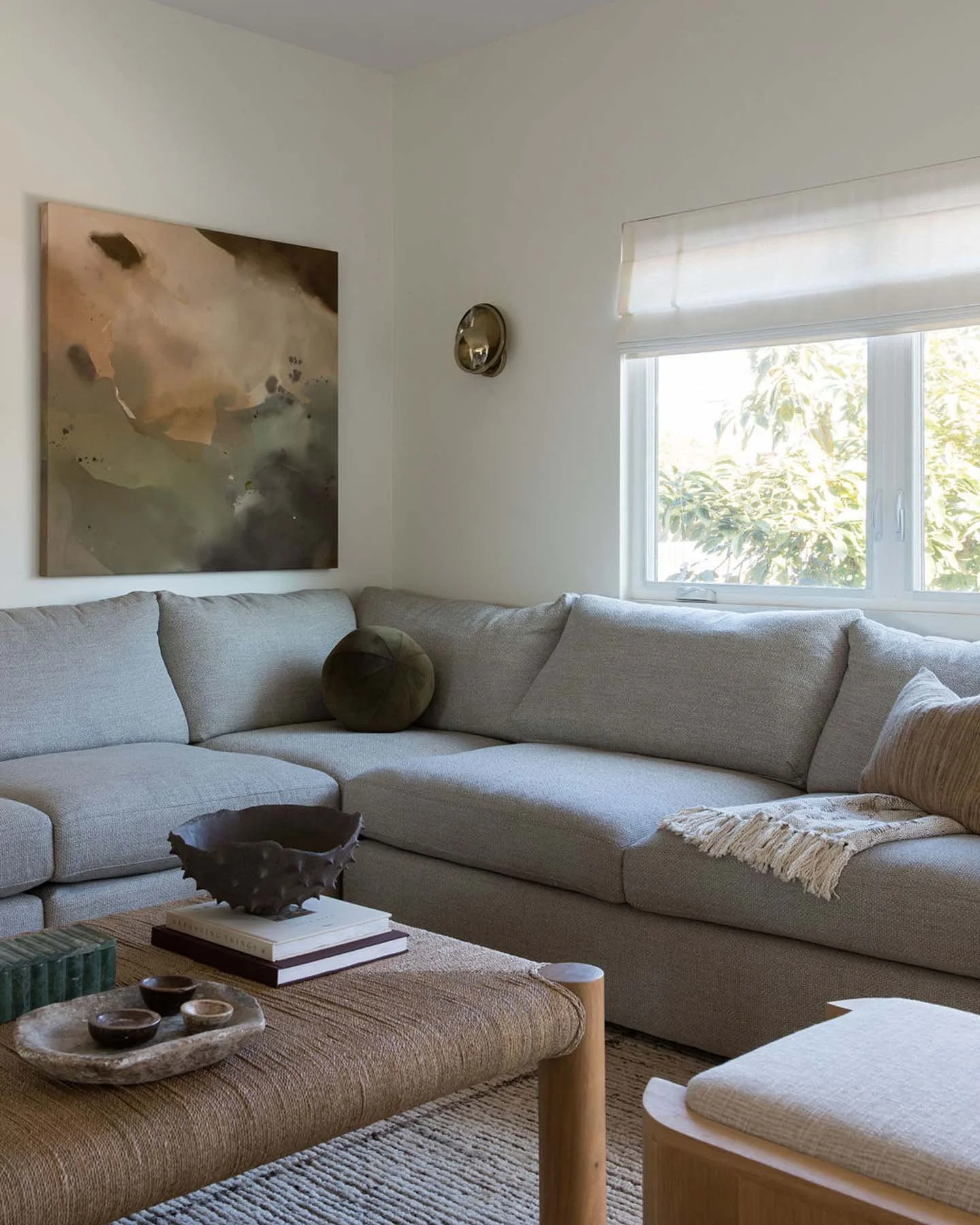 Living room with beige sectional sofa, green pillows, abstract wall art, window with white shades, coffee table with decorative objects, and a beige rug.