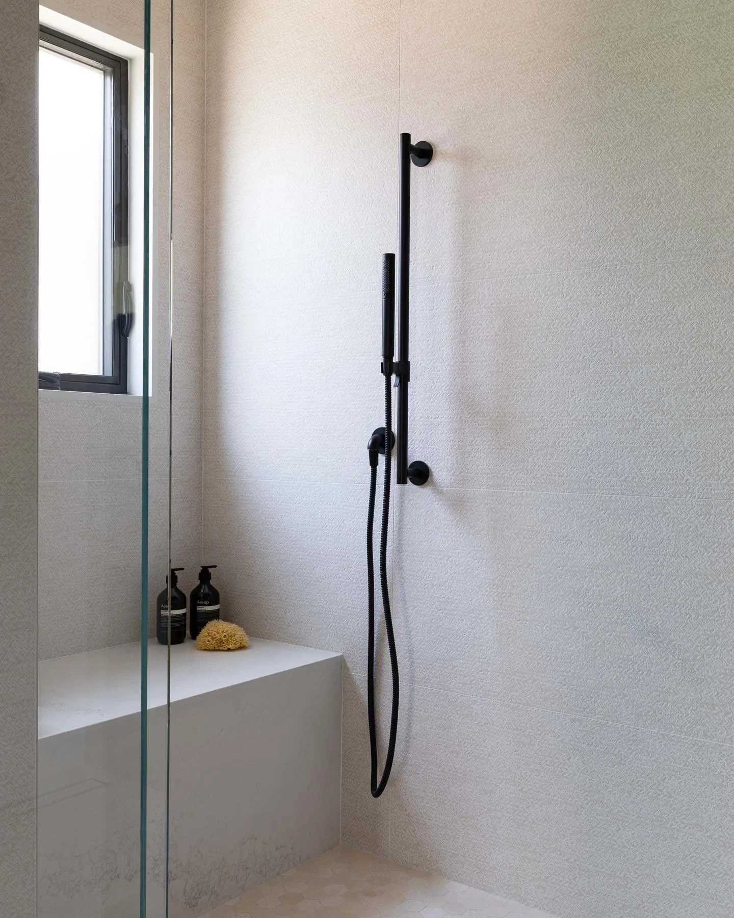 Modern shower area with a handheld showerhead attached to a black vertical bar on a beige textured wall. There is a small ledge with two bottles and a yellow bath sponge. A window is visible on the left, and a glass shower door is partially shown.