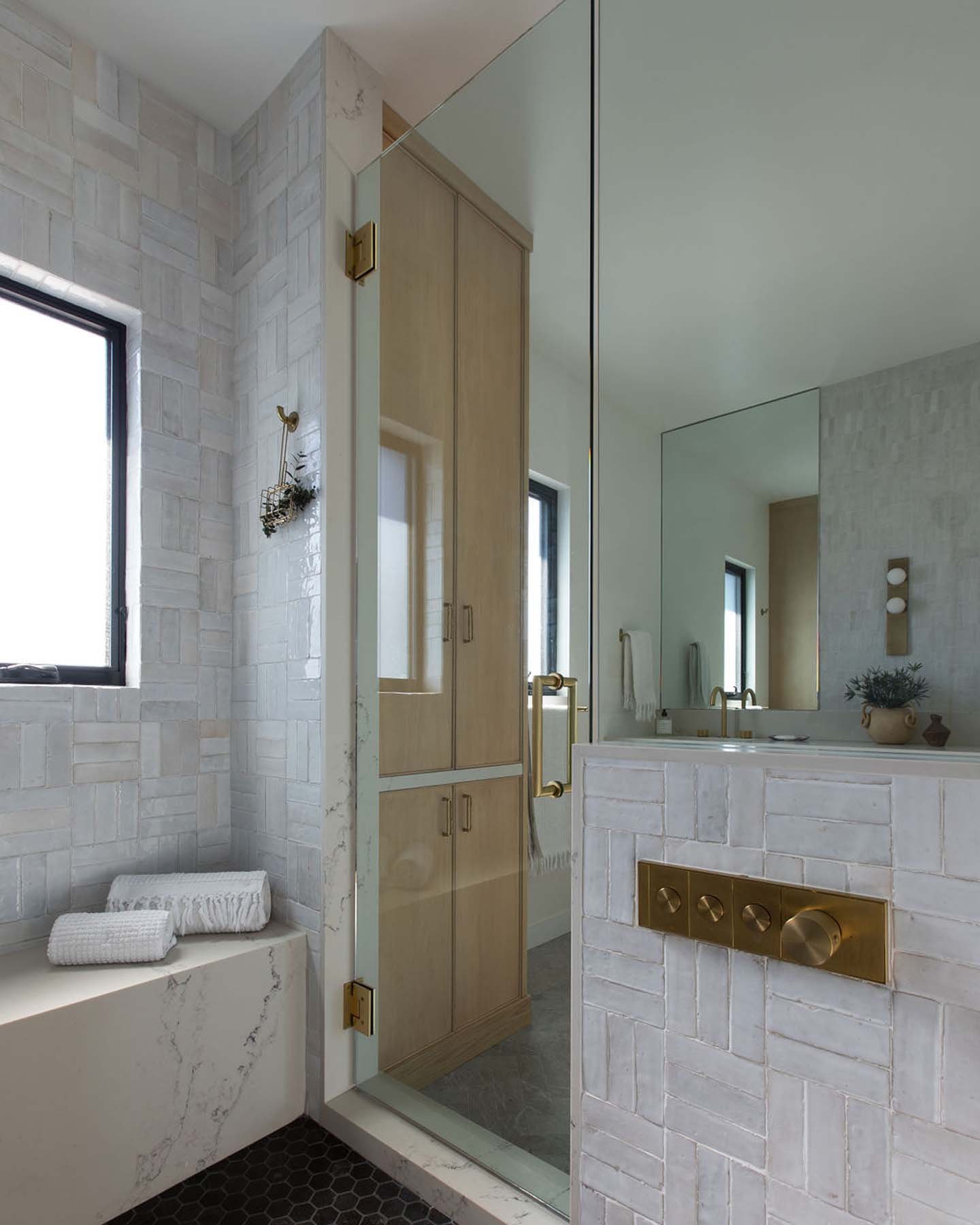 A modern bathroom with white brick tiles, a black hexagonal tile floor, a window, and a glass shower enclosure. There is a wooden cabinet, gold fixtures, a white bathtub with rolled towels, a mirror, and a plant on the vanity.