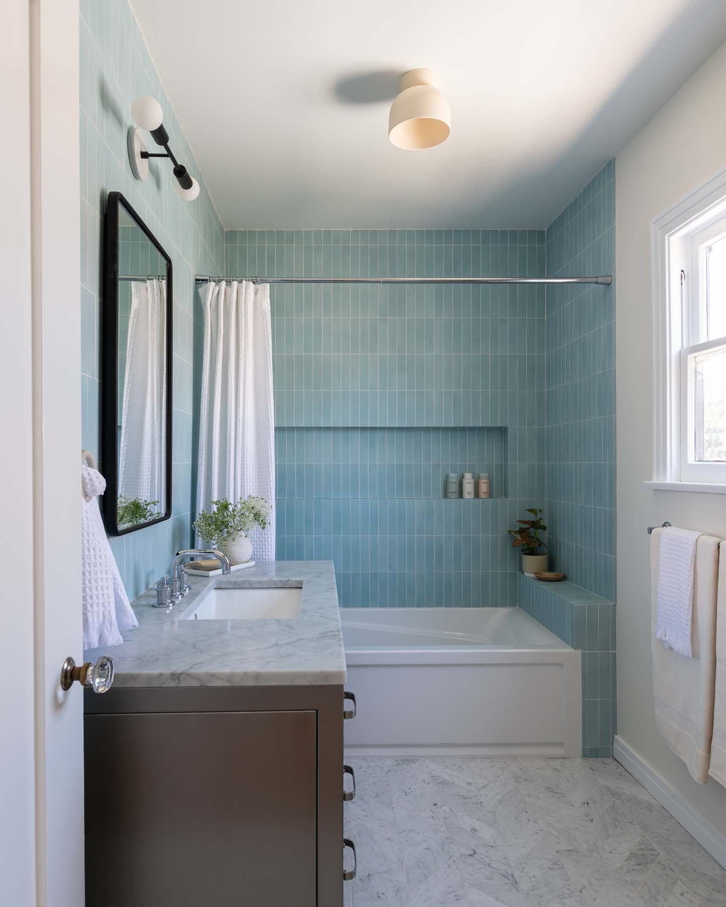 A modern bathroom with a light blue tiled shower area, a white bathtub, a marble sink countertop, a mirror, a window, and minimal decor including potted plants and white towels.