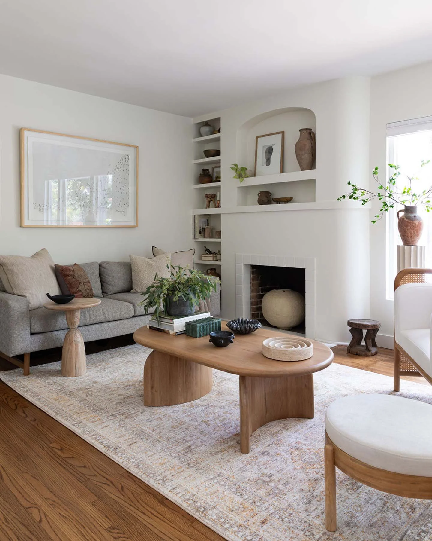 Living room with gray sofa, wooden coffee table, beige rug, and decorative vases on shelves and a window with greenery.