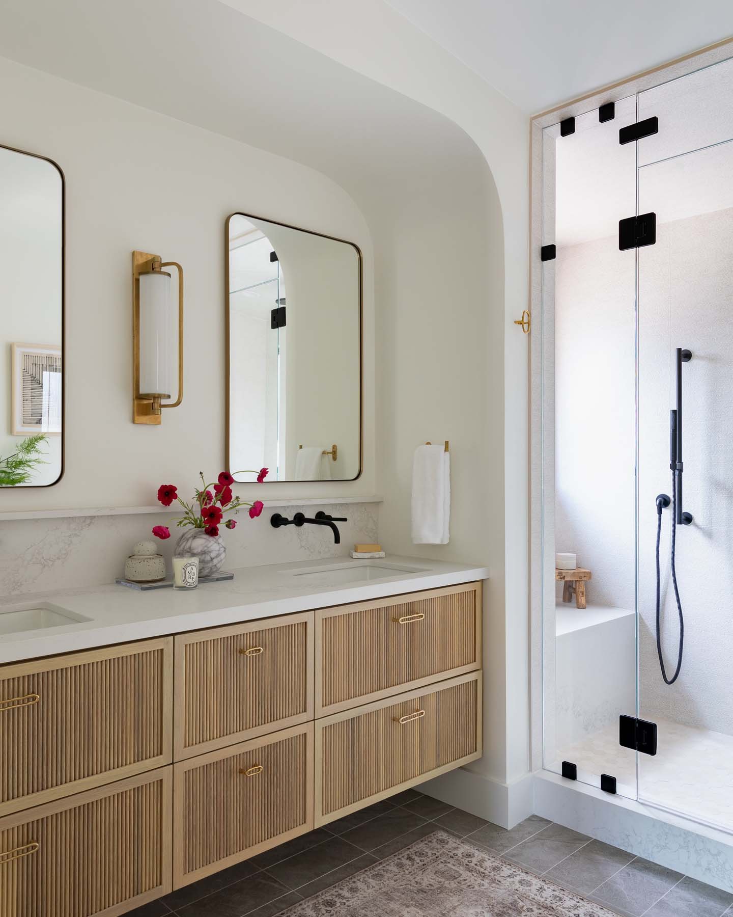 A modern bathroom with a beige vanity with vertical grooves, four drawers, and a white countertop with a circular sink. Above the vanity are two rectangular mirrors and a brass wall-mounted light fixture. To the right, there is a walk-in shower with a glass door, a black showerhead, and fixtures. The shower has a small wooden stool and a textured light-colored wall.
