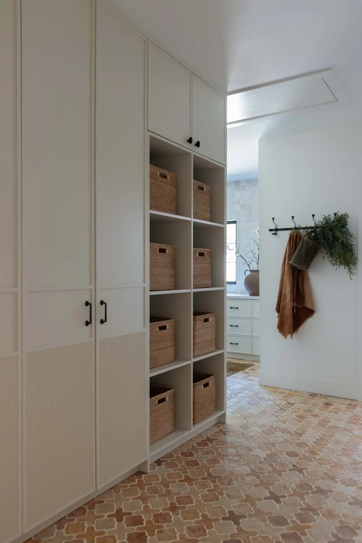 A closet area with built-in white cabinets, woven baskets on open shelves, and a wall hook with hanging brown and green textiles, with a window and decorative vase in the background.