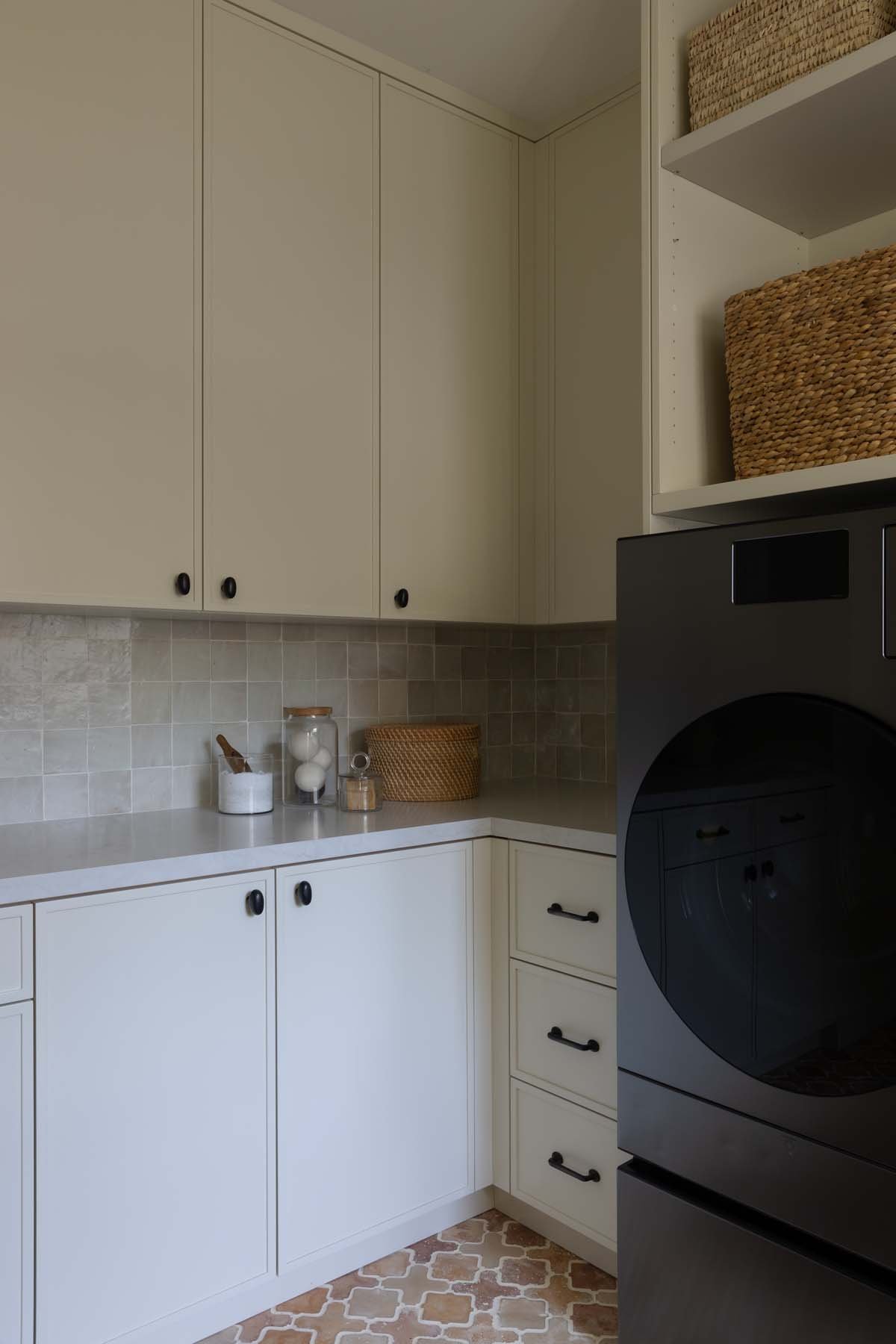 A corner of a kitchen with white cabinets, a beige tiled backsplash, and a black washing machine. Decor includes glass jars, a woven basket, and small containers on the countertop.