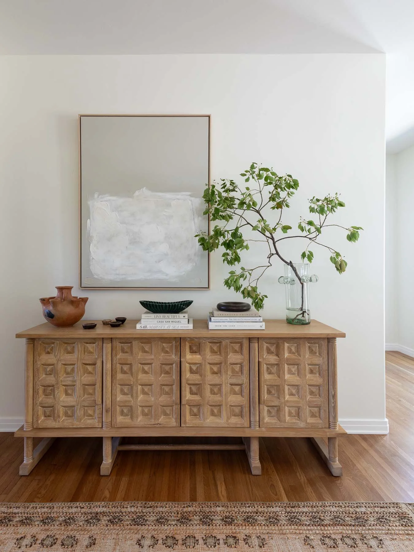 A wooden sideboard with geometric carved doors on a hardwood floor. Decor includes ceramic and glass vases, bowls, and stacked books. A large mirror hangs on a white wall, and a leafy plant in a glass vase is on the right side of the sideboard. Part of a patterned rug is visible at the bottom.