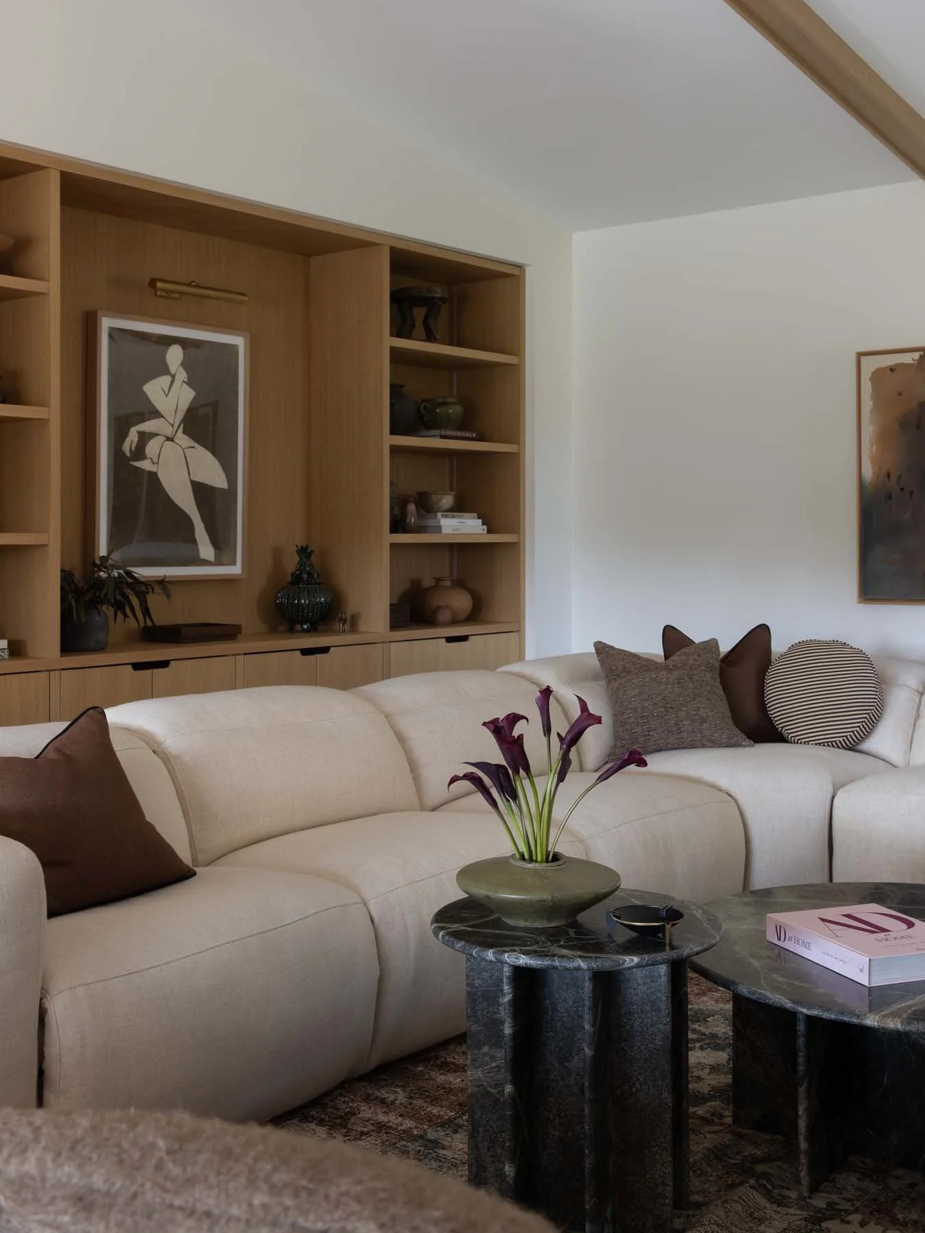 Living room with beige sofa, decorative pillows, dark patterned rug, black marble tables, a bouquet of dark purple calla lilies in a gray vase, wooden shelving with art and decor, and a painting on the white wall.
