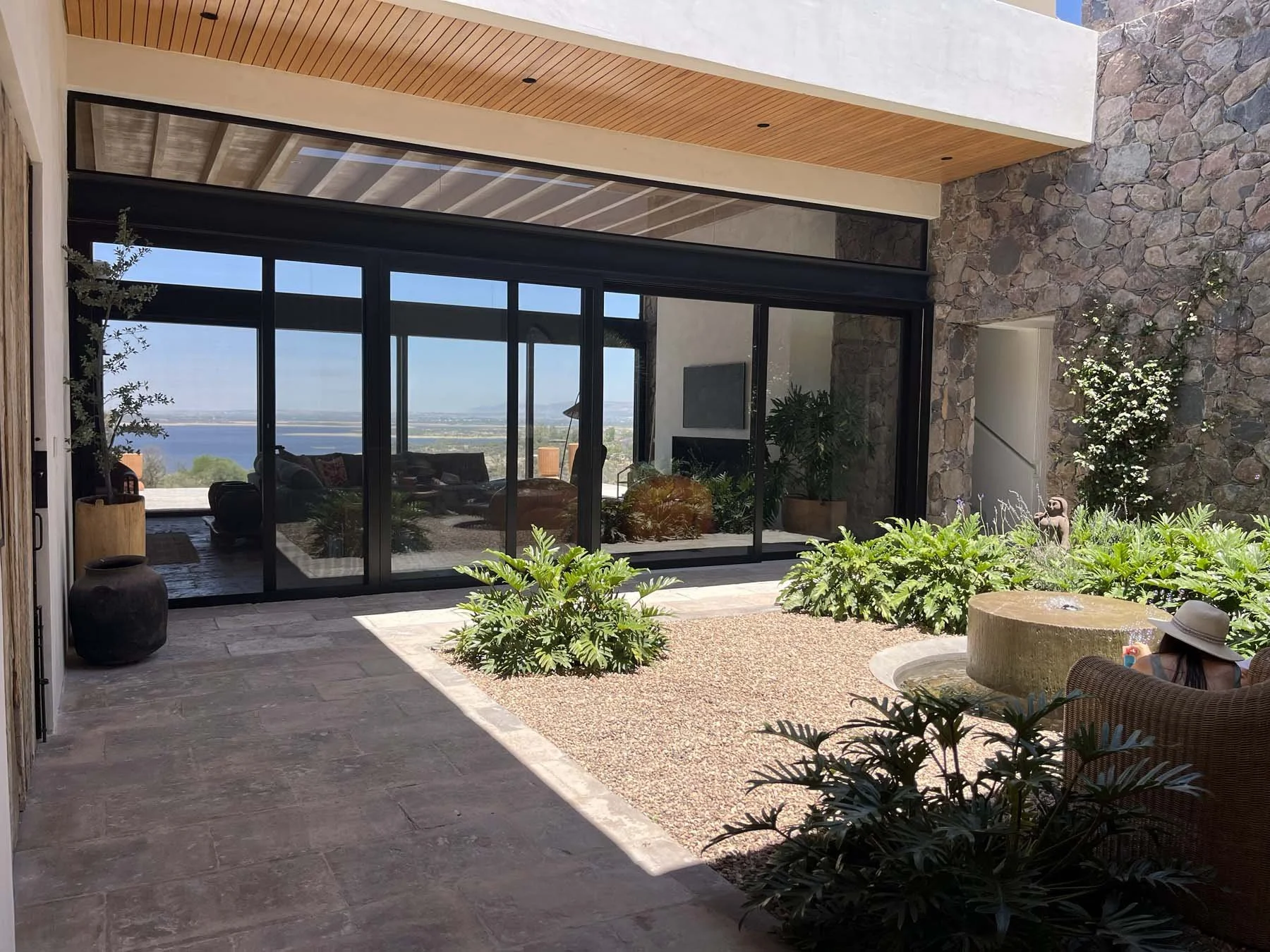 Modern outdoor patio with sliding glass doors, stone wall, and lush greenery, overlooking a body of water in the distance.
