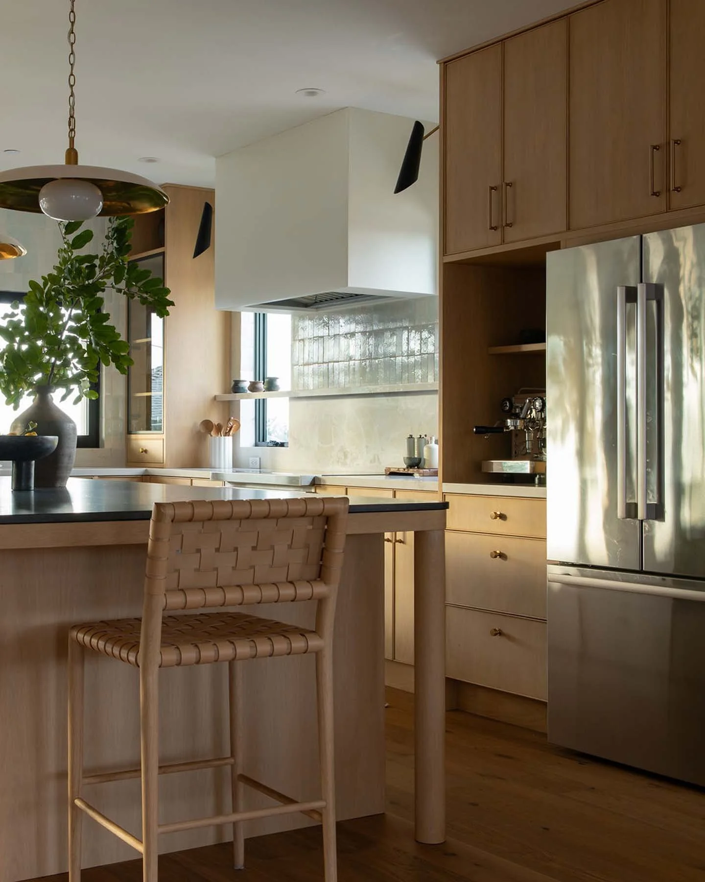 Modern kitchen interior with wooden cabinets, stainless steel refrigerator, and a kitchen island with a woven chair, green plants, and decorative items.