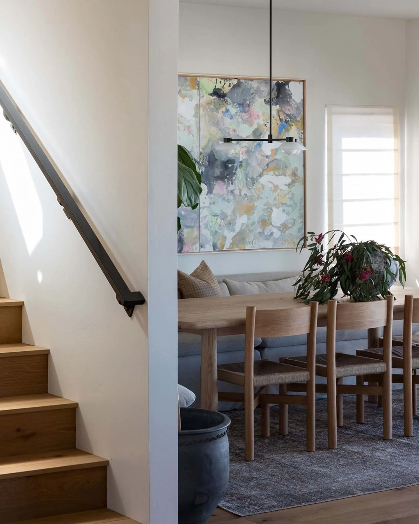 Interior of a home showing a wooden dining table with chairs, a large leafy plant, an abstract painting on the wall, and a window with white blinds, with carpeted flooring.