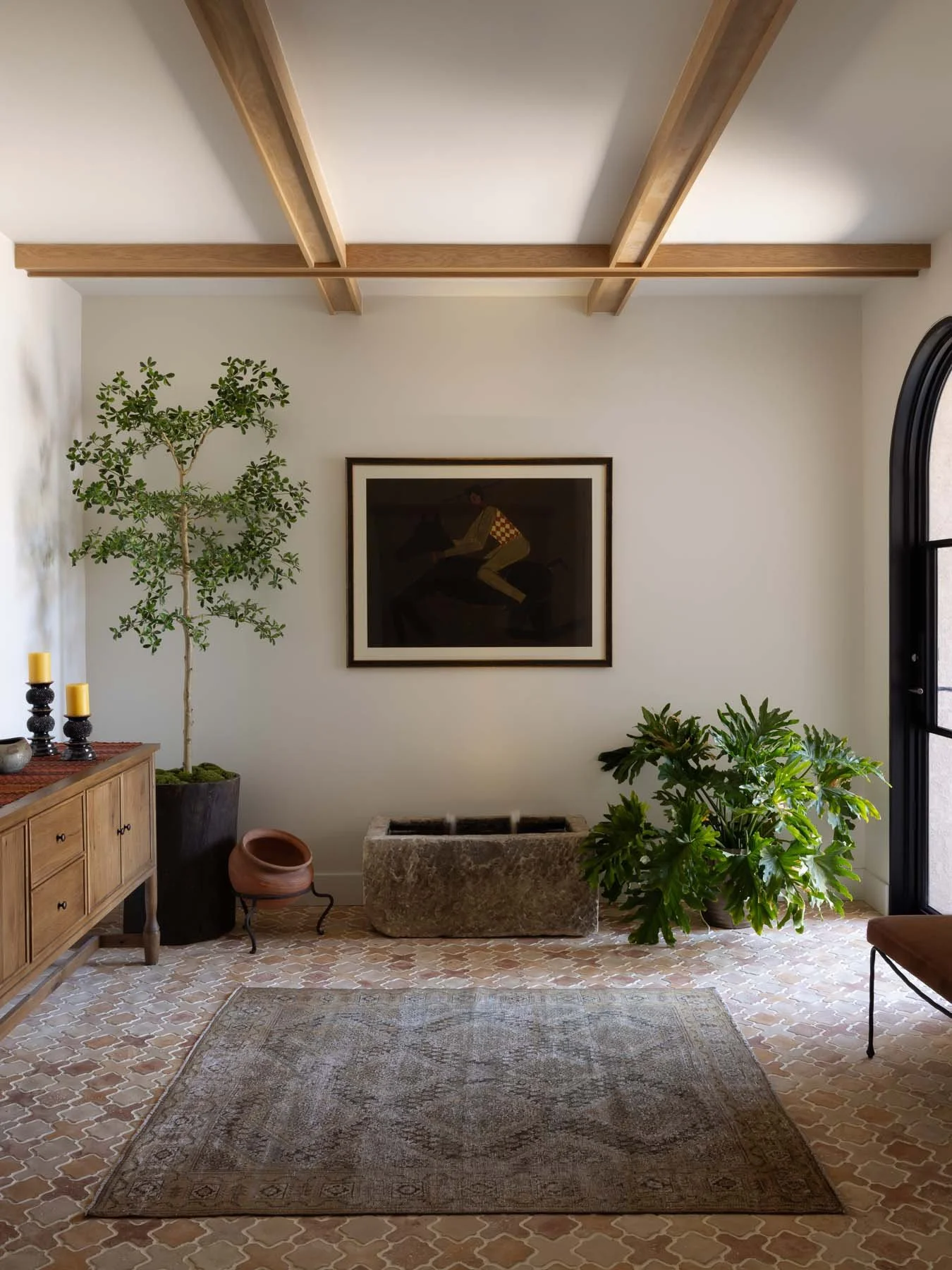 Living room with beige tiled floor, area rug, green potted plants, a wooden sideboard with candles, framed artwork, and a large stone planter.