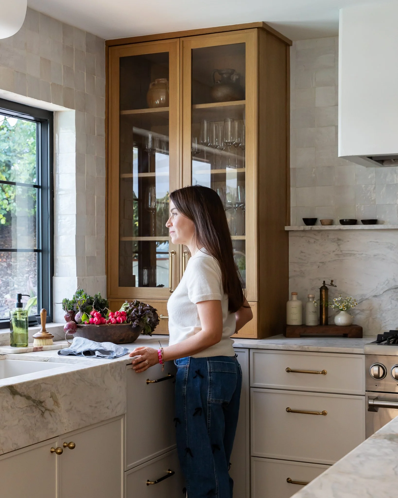 A woman with long brown hair, dressed in a white shirt and jeans, standing in a kitchen with white cabinets, a marble countertop, and a large window with natural light. She is near a wooden bowl of fresh radishes and vegetables.