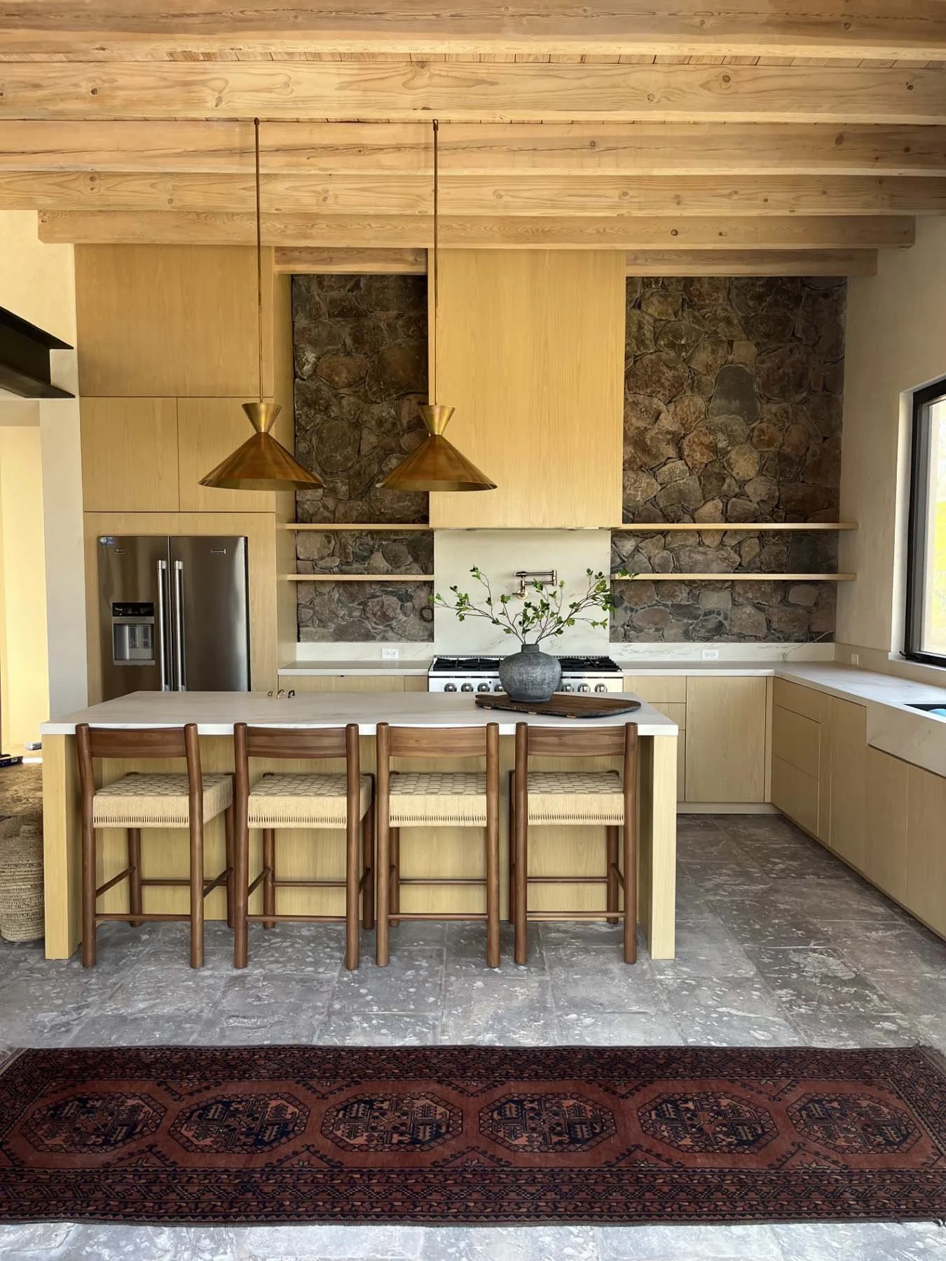 Modern kitchen with wood cabinets, stone backsplash, a kitchen island with four wooden chairs, hanging pendant lights, a window on the right, and a red patterned rug on the floor.