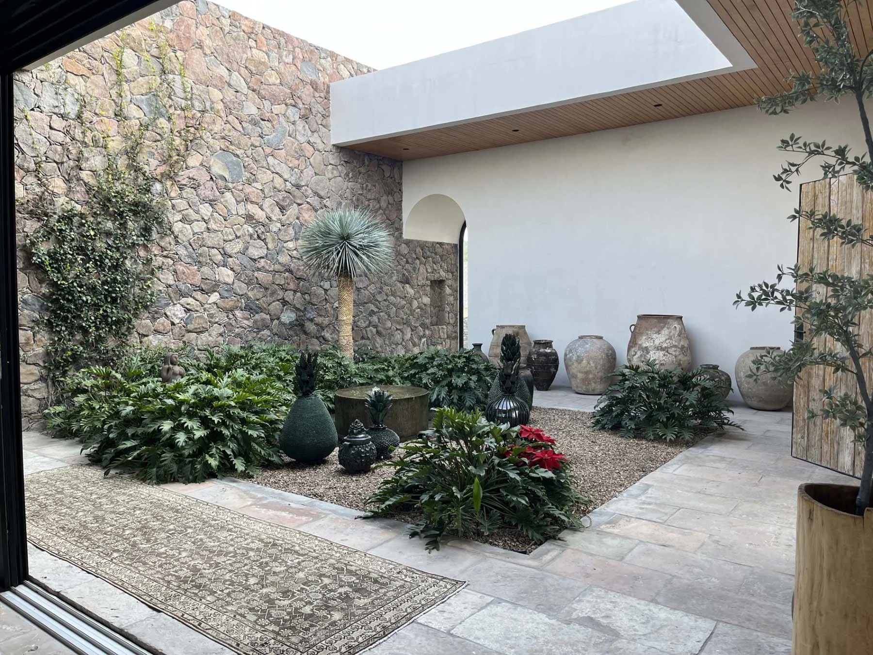 Indoor-outdoor courtyard featuring a stone wall, various potted plants, decorative jars, and a large rug on a light-colored tiled floor.
