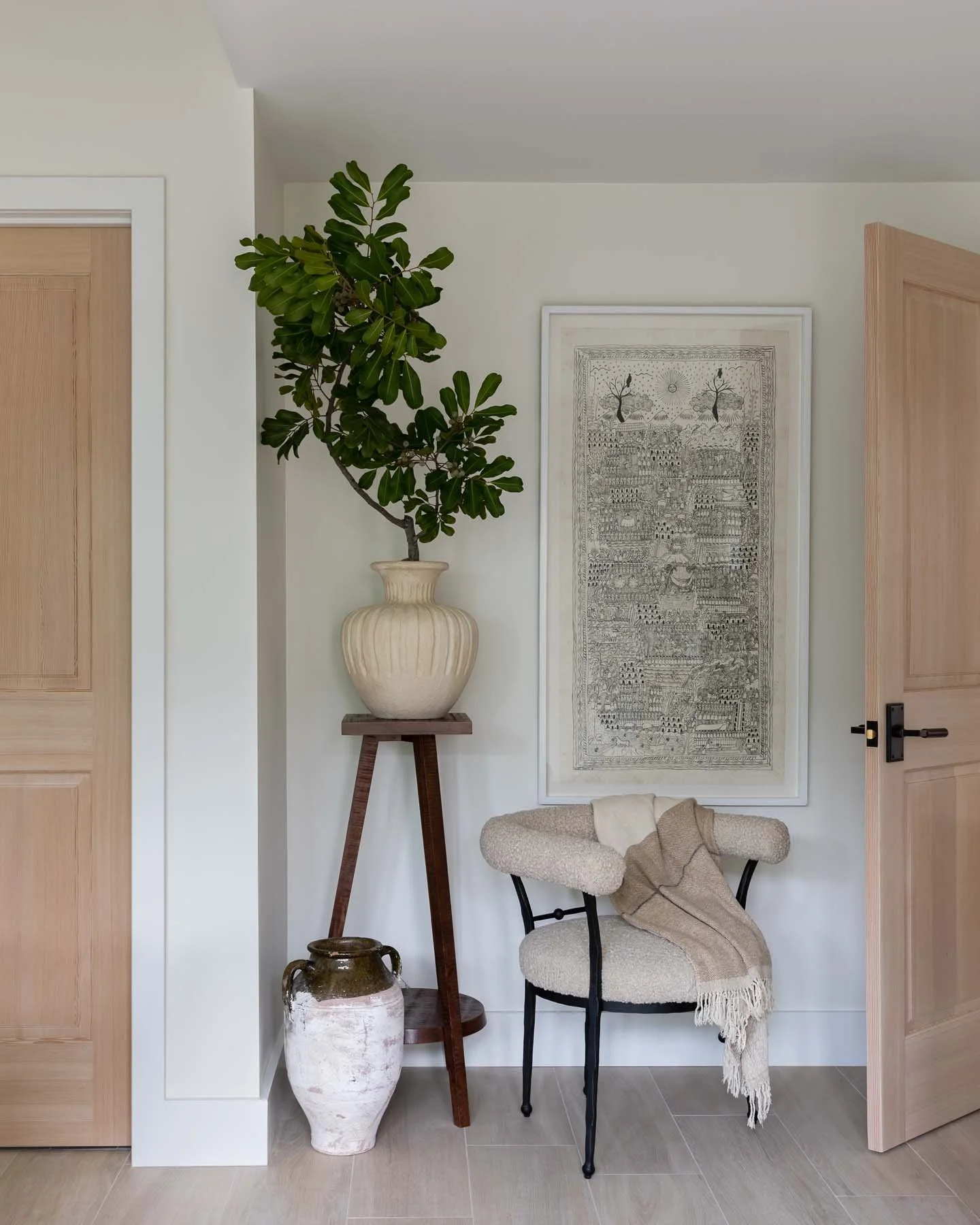 Interior decorative corner with a potted plant on a stand, a framed art piece on the wall, a woven chair with a beige throw, a large vase on the floor, and centered wooden doors.