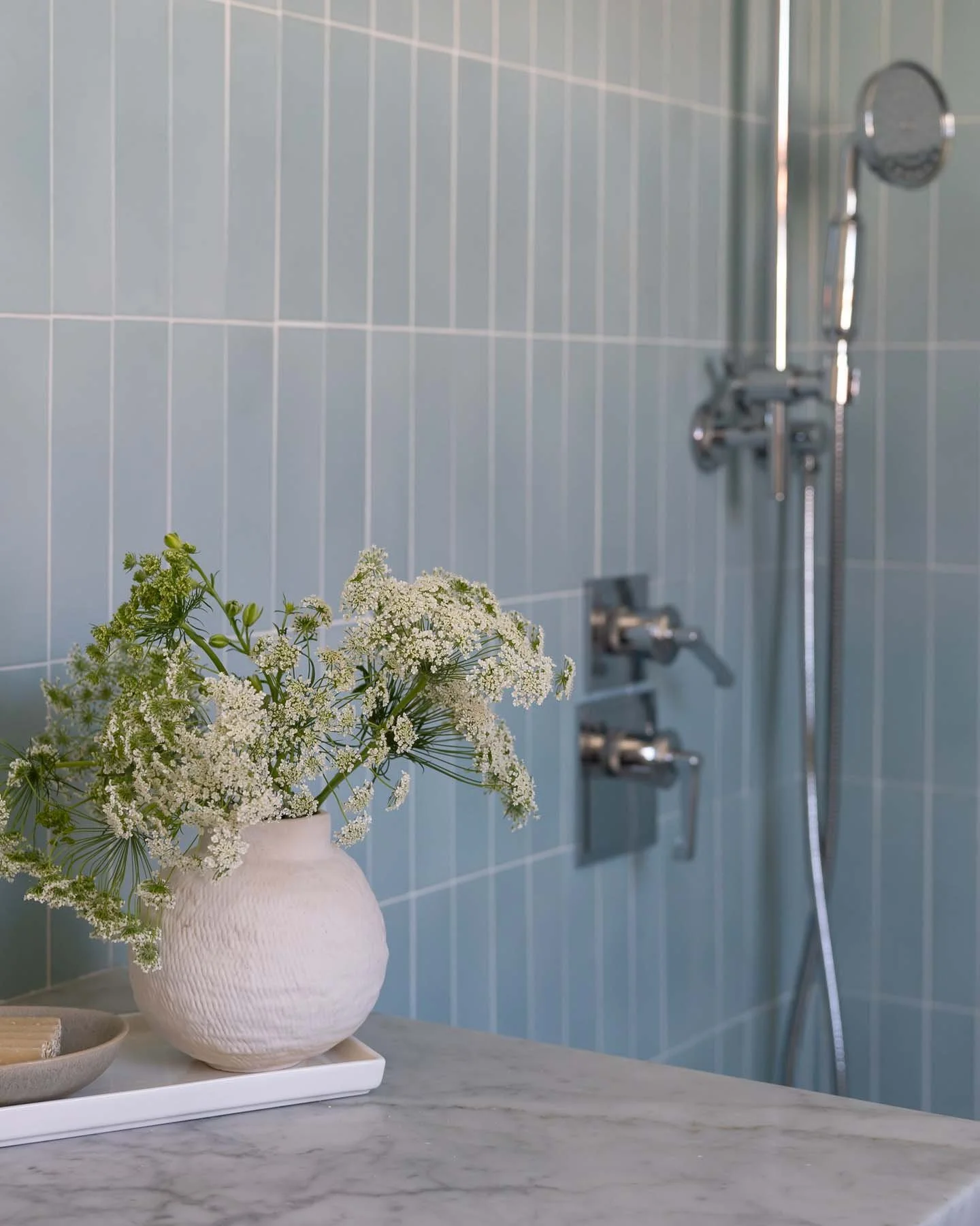 White ceramic vase with delicate white flowers and green foliage on a marble surface, with a blue tiled shower wall and silver shower fixtures in the background.