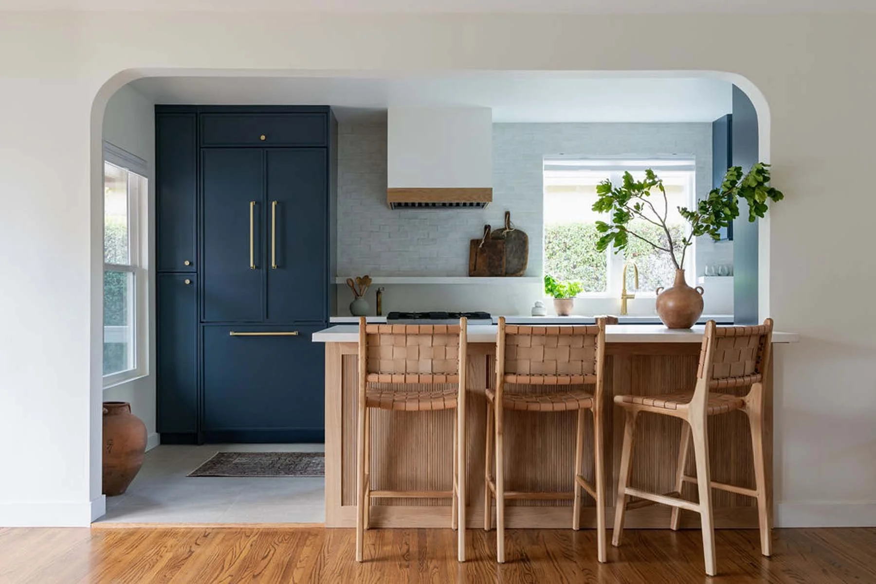 Modern kitchen with dark blue cabinets, a white range hood, a kitchen island with wooden chairs, large window with greenery outside, and decorative vases and plants on the countertop.