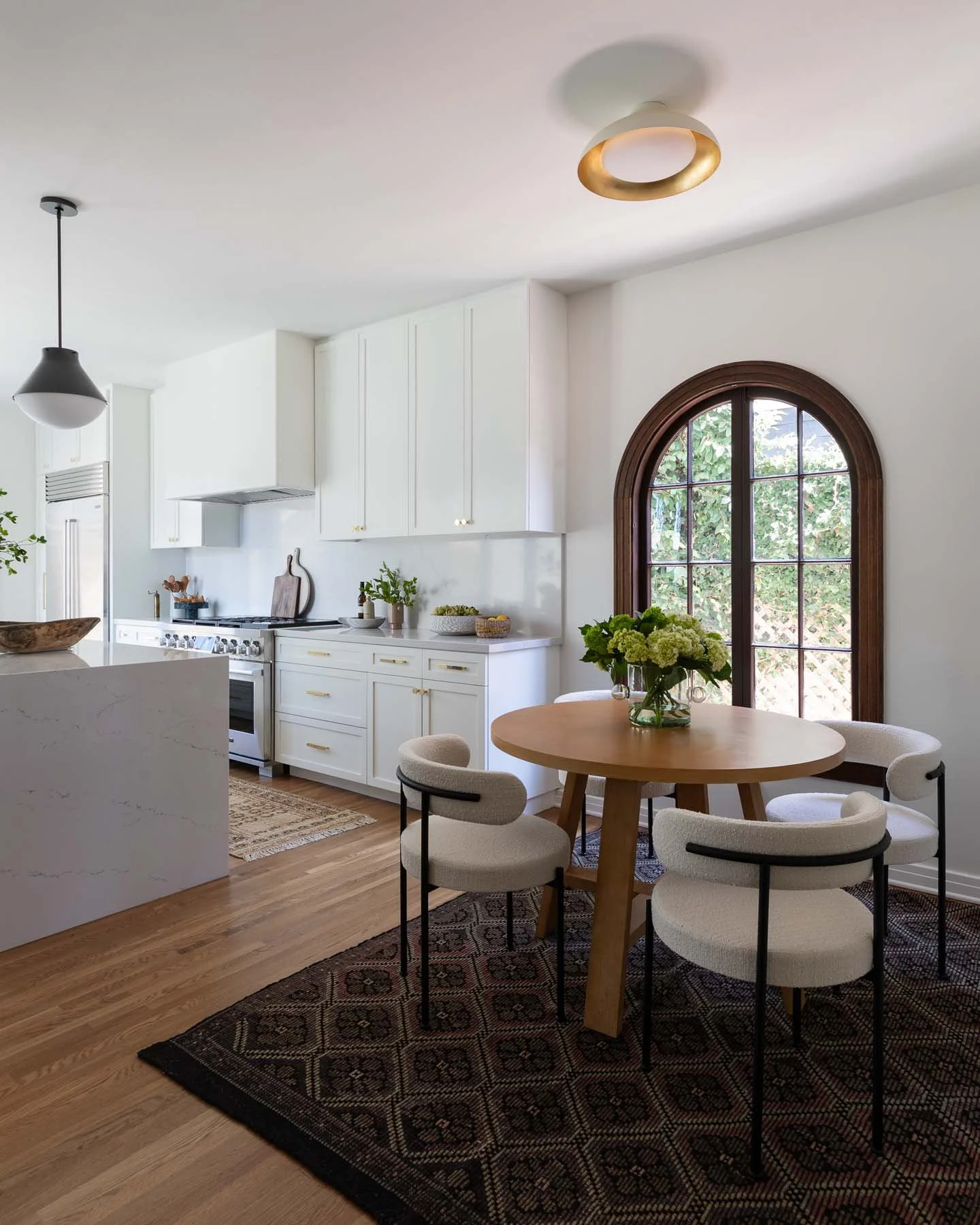 Modern kitchen and dining area with white cabinets, a round wooden table, six upholstered chairs, a window with wooden trim, and a patterned rug.