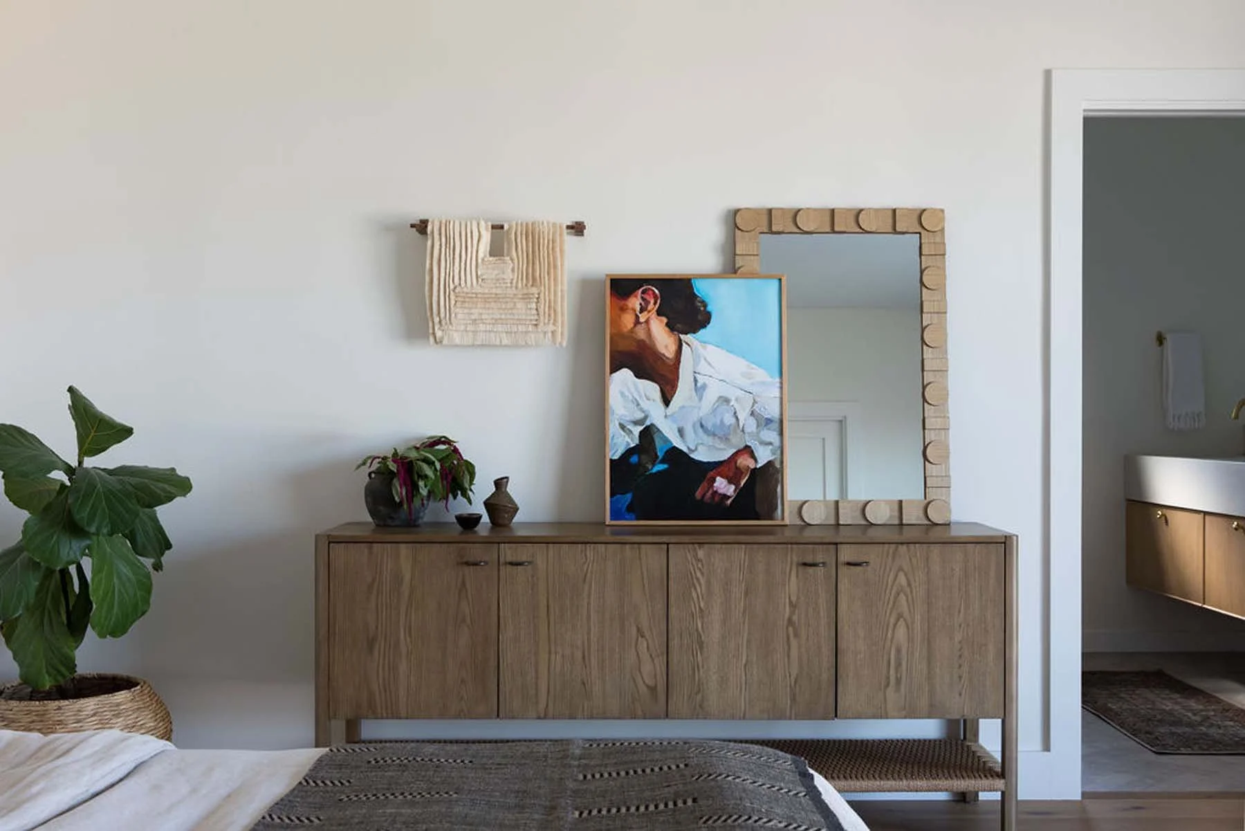 A wooden sideboard with a large framed painting of a woman sitting, a mirror with a wooden frame, and decorative vases with plants on top. A wall hanging and a houseplant are also visible.
