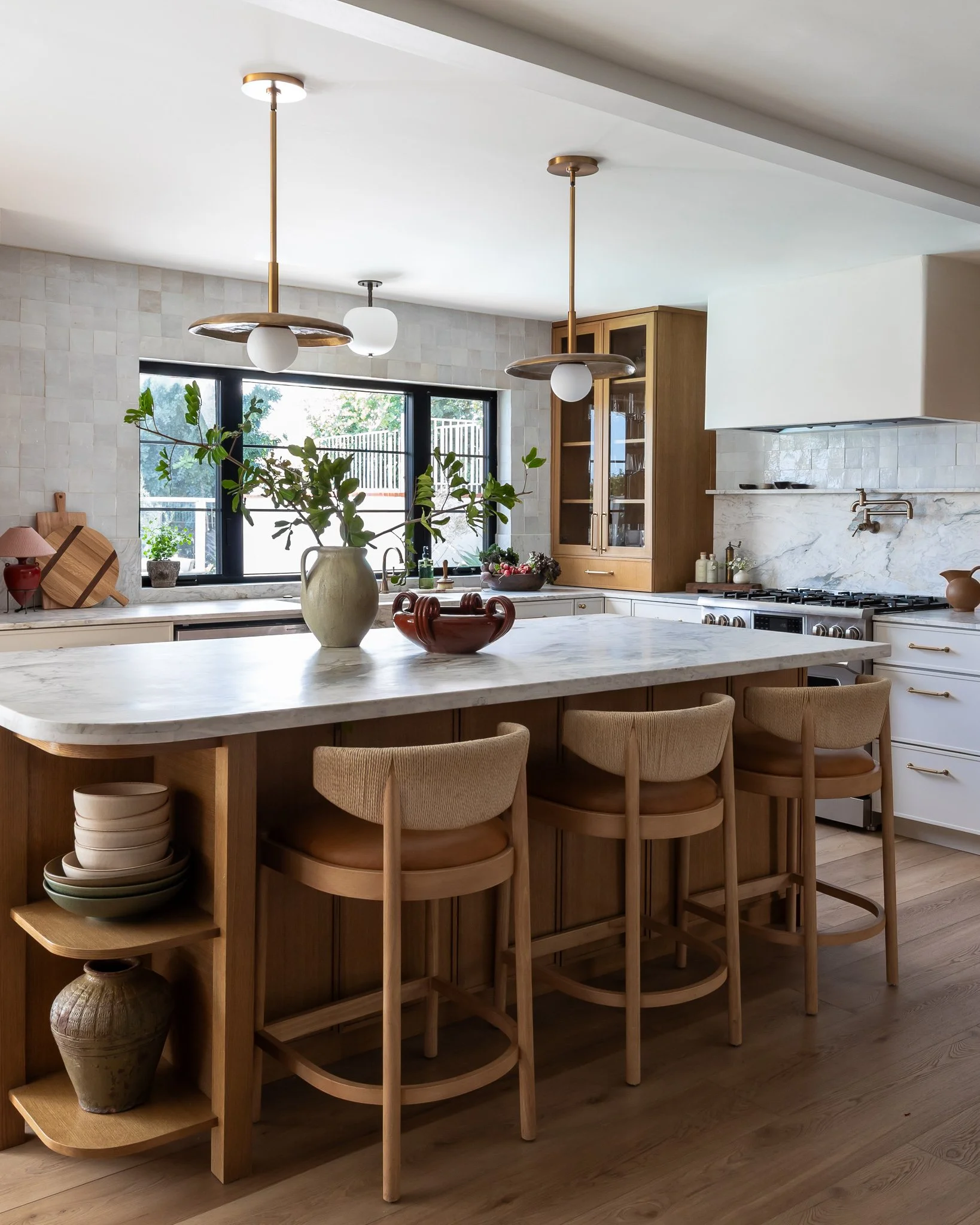 A modern kitchen with a white marble island, beige bar stools, a window with a view of trees, and wooden cabinets.