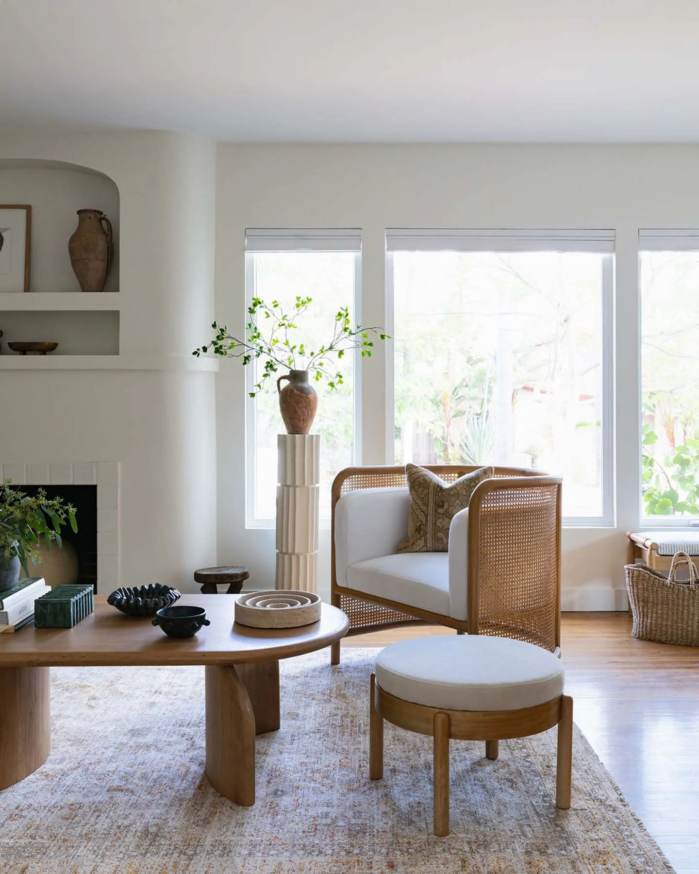 Bright living room with large windows, white walls, minimalist furniture, a wooden coffee table with decorative objects, a white armchair with a rattan frame, a round ottoman, a tall vase with green branches, and a built-in shelf with pottery.