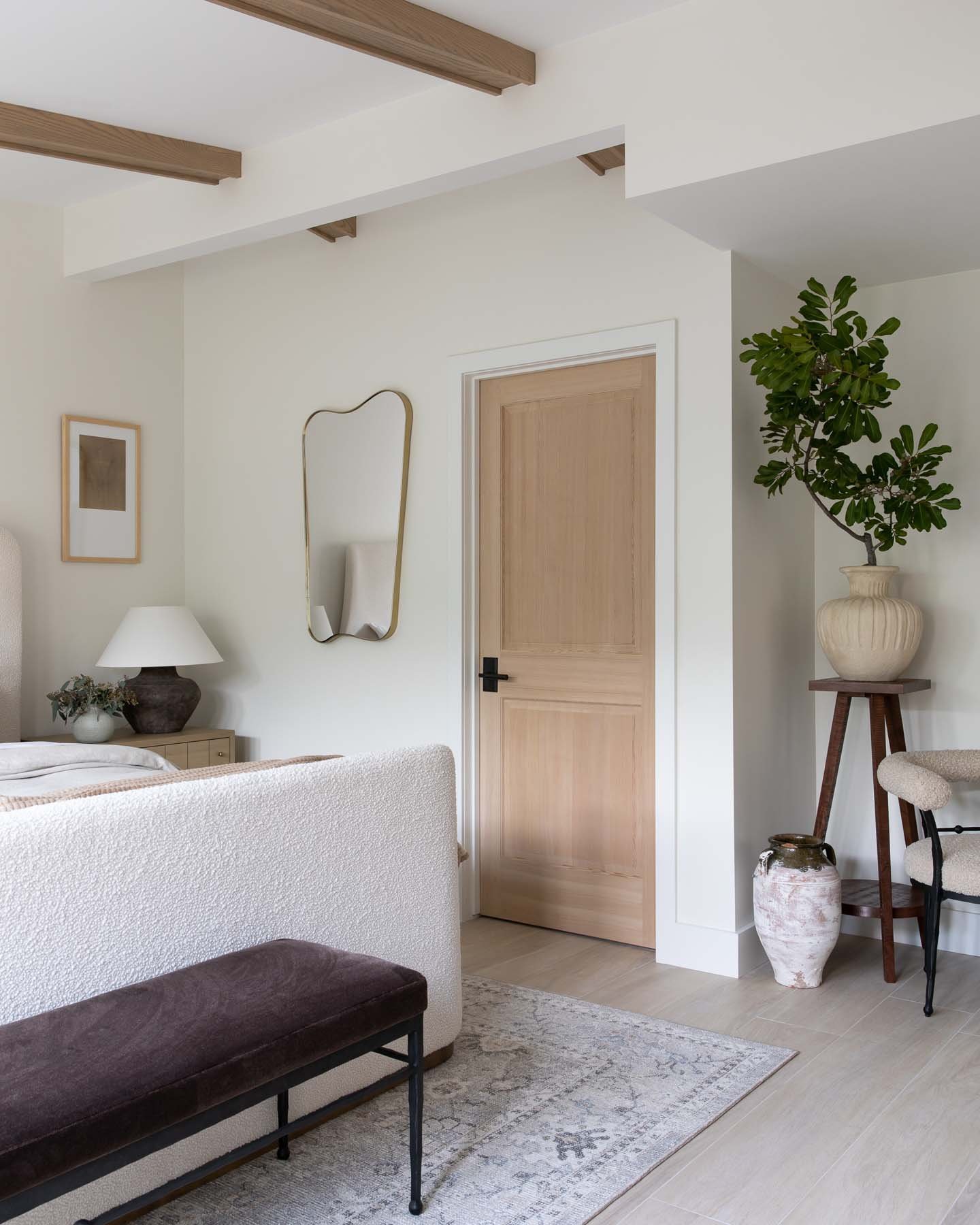 A cozy living room corner featuring a beige upholstered sofa, a dark wood bench, a side table with a white lamp and decorative plant, a tall potted plant on a stand, a mirror, and a wooden door in the background, all with neutral tones and minimal decor.