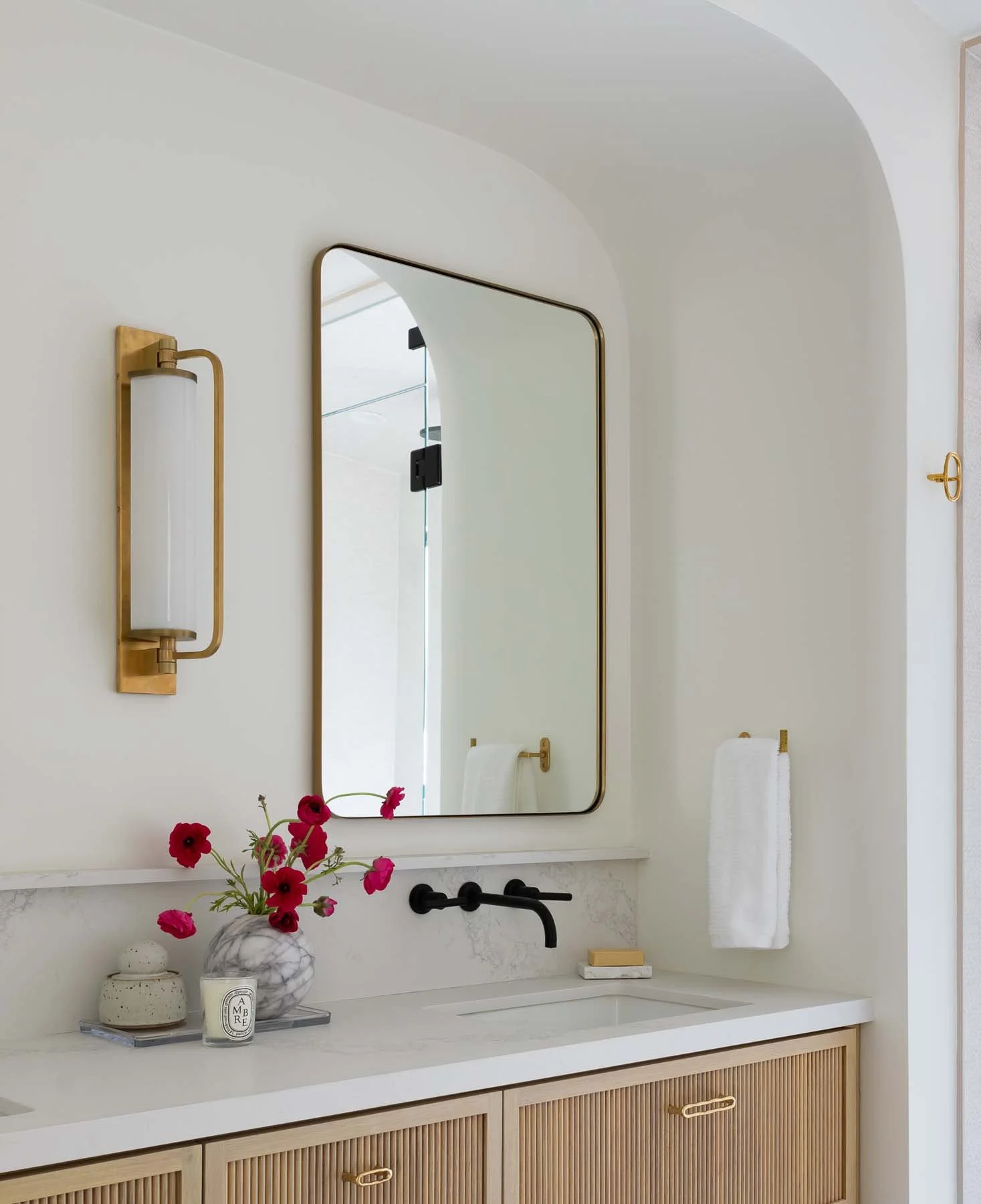 Bathroom vanity area with a large mirror, a black faucet, and white countertop. Decor includes pink flowers in a marble vase, a small candle, and a soap dish. Gold light fixture, towel, and cabinet handles.