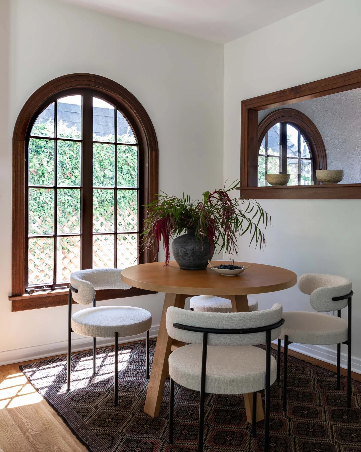 A dining room featuring a round wooden table with five cream-colored chairs. A dark gray vase with green and red foliage and a small bowl are on the table. There are large arched windows with wooden trim allowing natural light. A mirror on the wall reflects the window, with two stone bowls on the window frame.