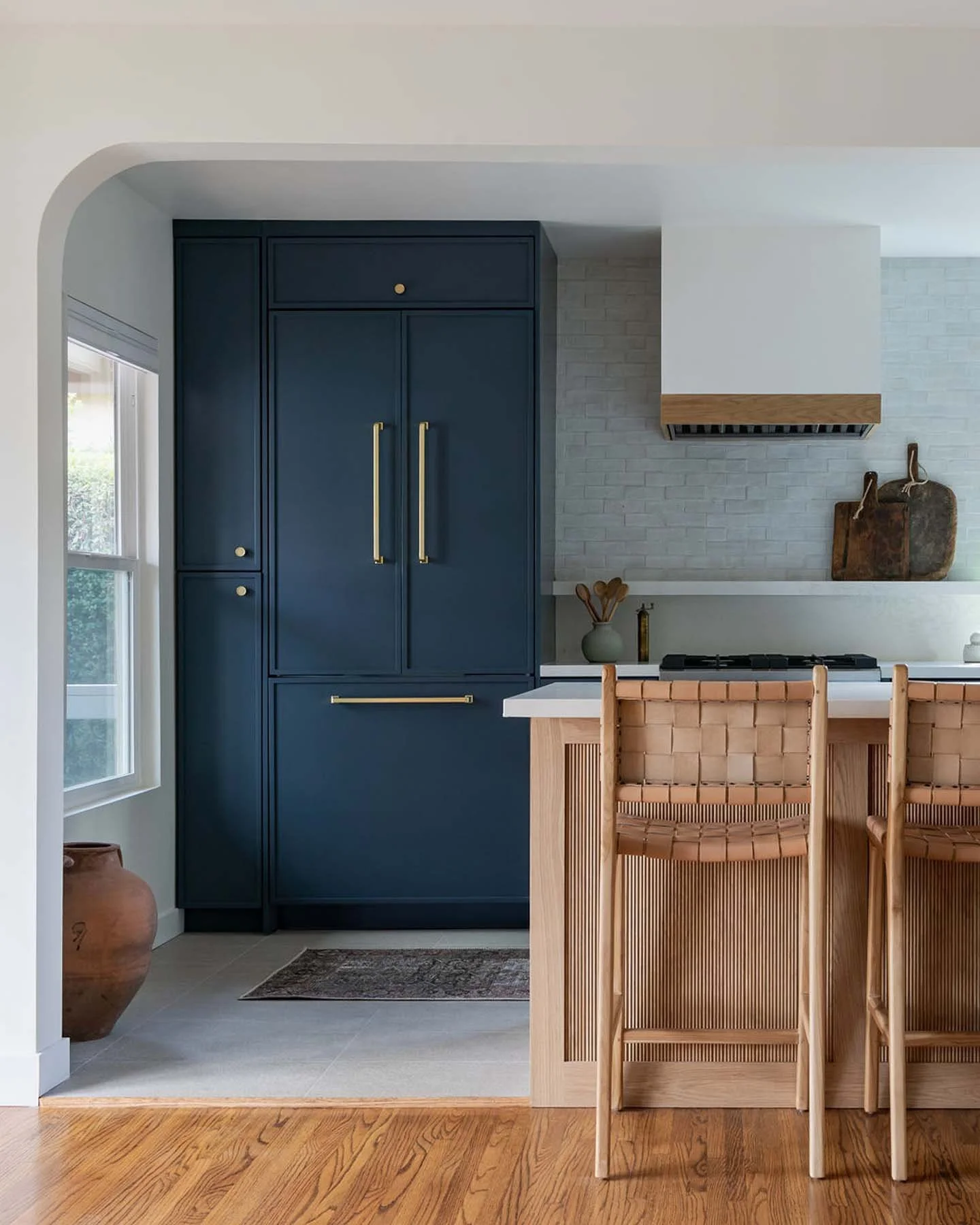 Modern kitchen with navy blue cabinets, gold handles, white brick backsplash, and a light wood breakfast bar with woven chairs.