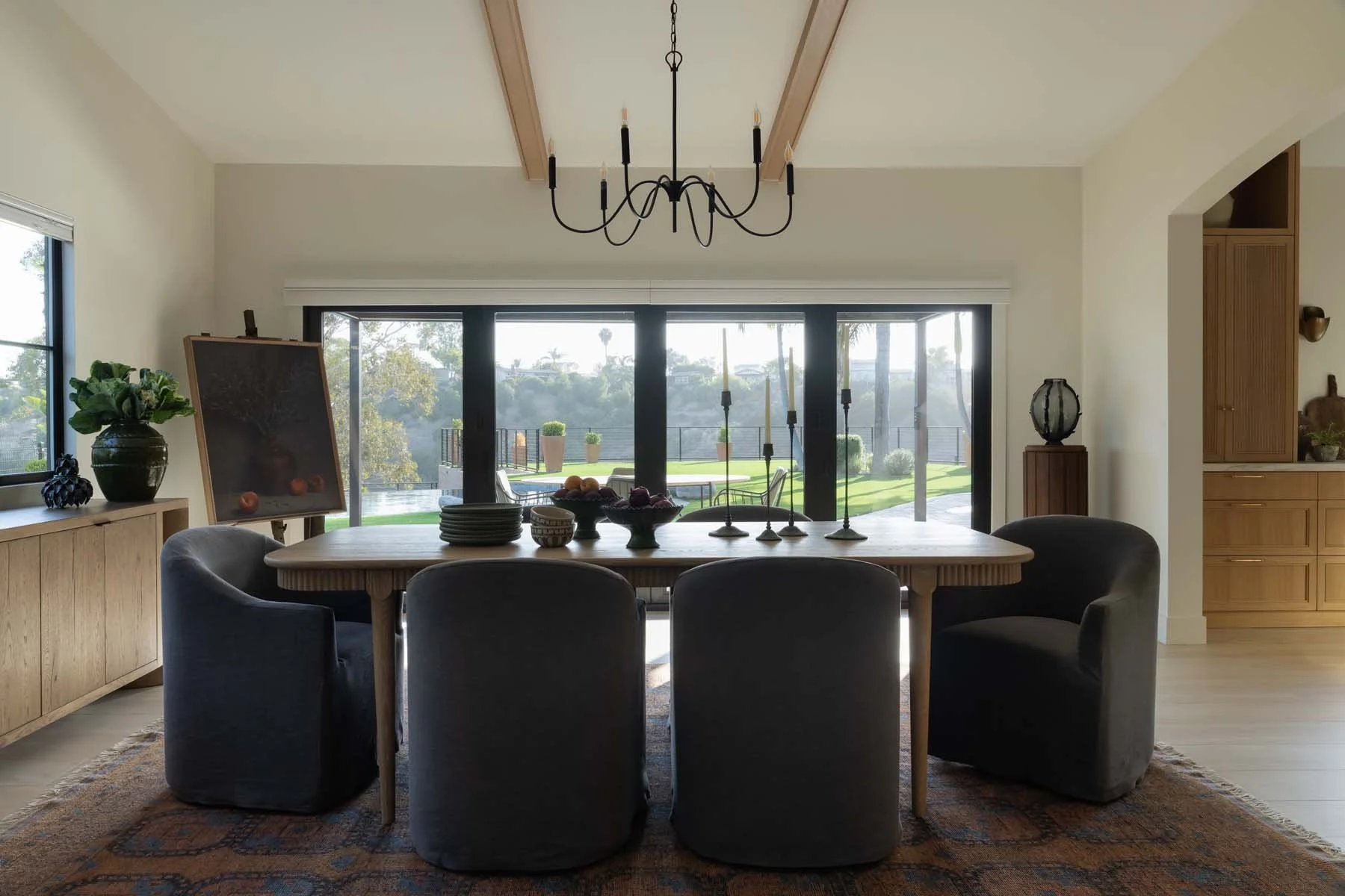 A dining room with a large wooden table and six dark upholstered chairs, decorative plates and bowls on the table, and a black chandelier hanging from the ceiling. Large sliding glass doors open to an outdoor patio with a pool and greenery. To the left, a sideboard with a potted plant and a painting on an easel. To the right, wooden cabinets and sculptures.