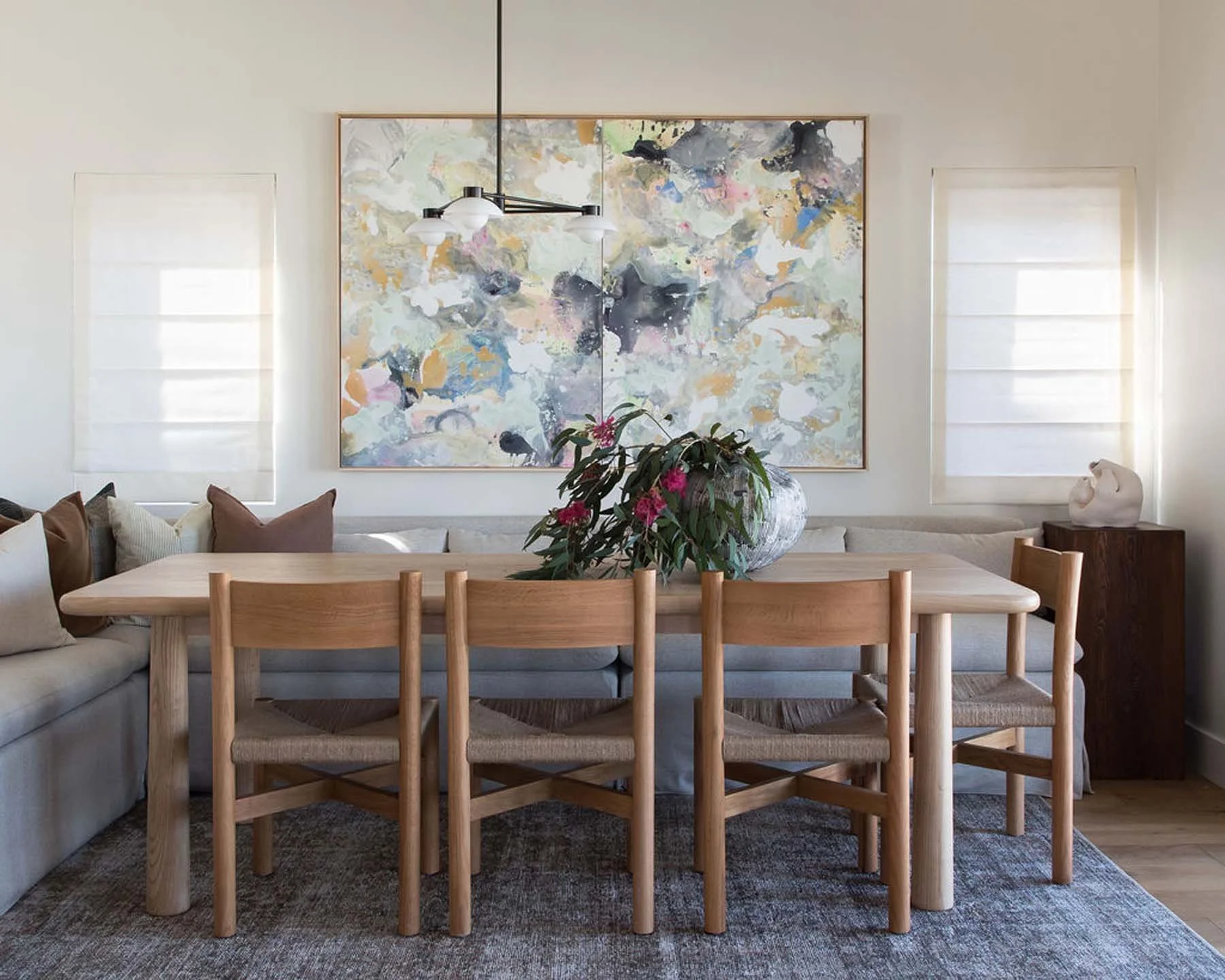 A dining room with a wooden table and chairs, a sofa with pillows, a large abstract artwork on the wall, a hanging light fixture, and a vase with pink flowers on the table.