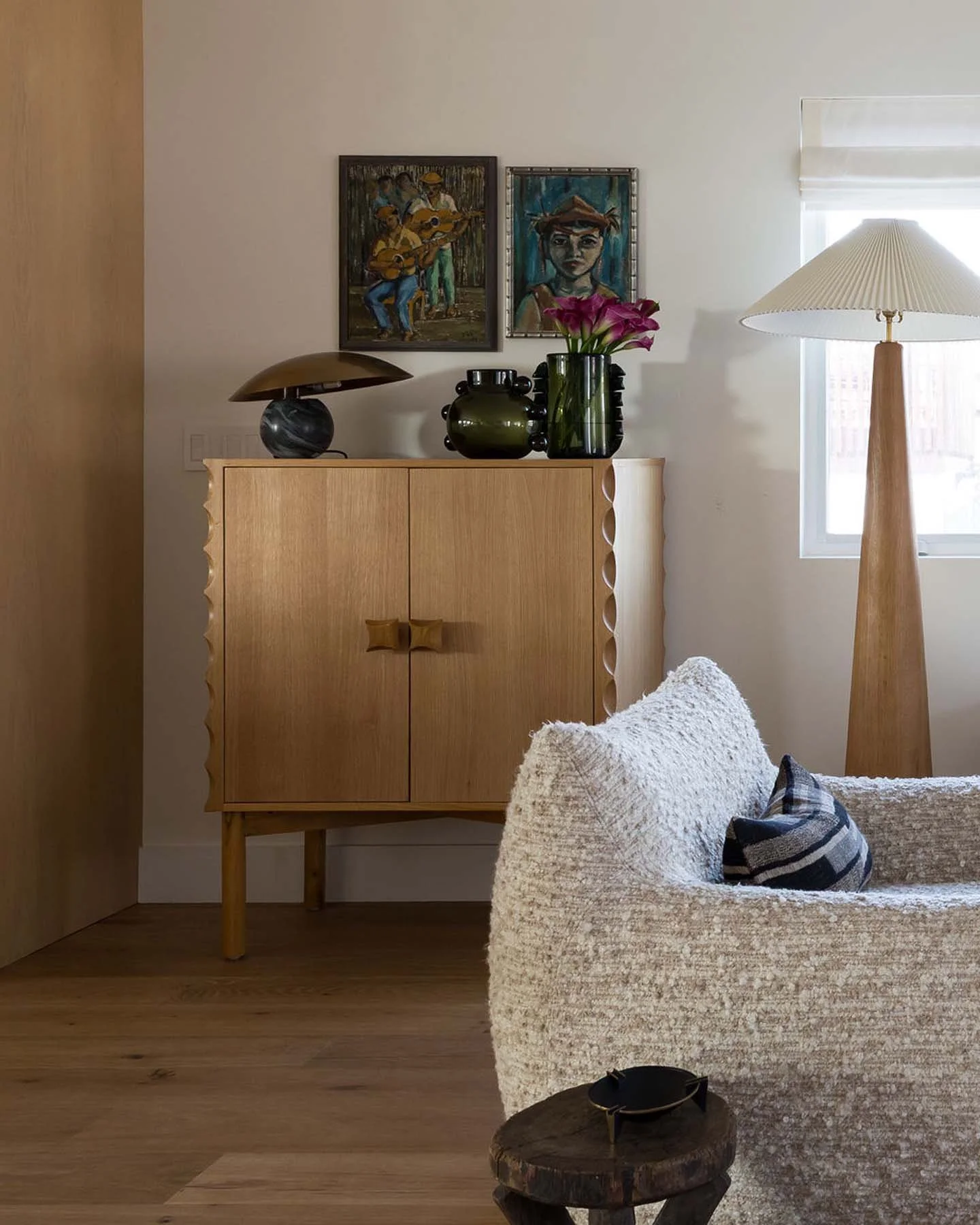 Living room with wooden cabinet, two paintings on wall, pink flowers in vase on cabinet, lamp, window with blinds, plush white couch with plaid pillow, small wooden side table with black bowl.